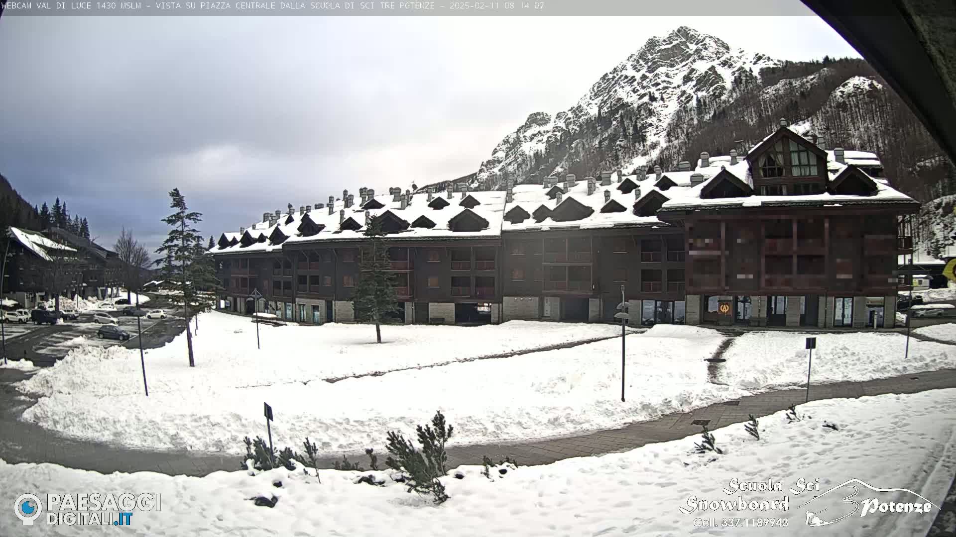 A snow-covered plaza sits in front of a large, multi-story building in a mountainous region under an overcast sky.