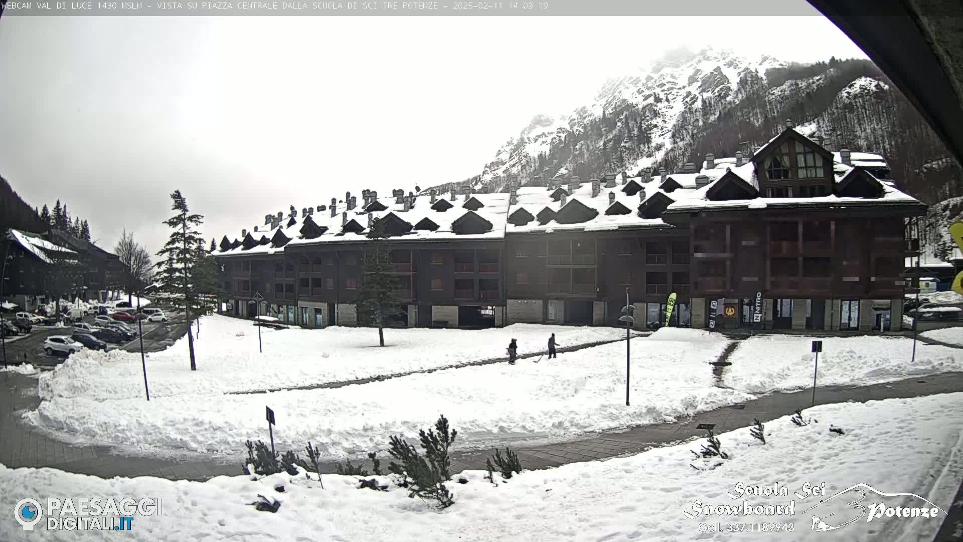 A snow-covered plaza with a large multi-story building sits before a mountain range under an overcast sky.