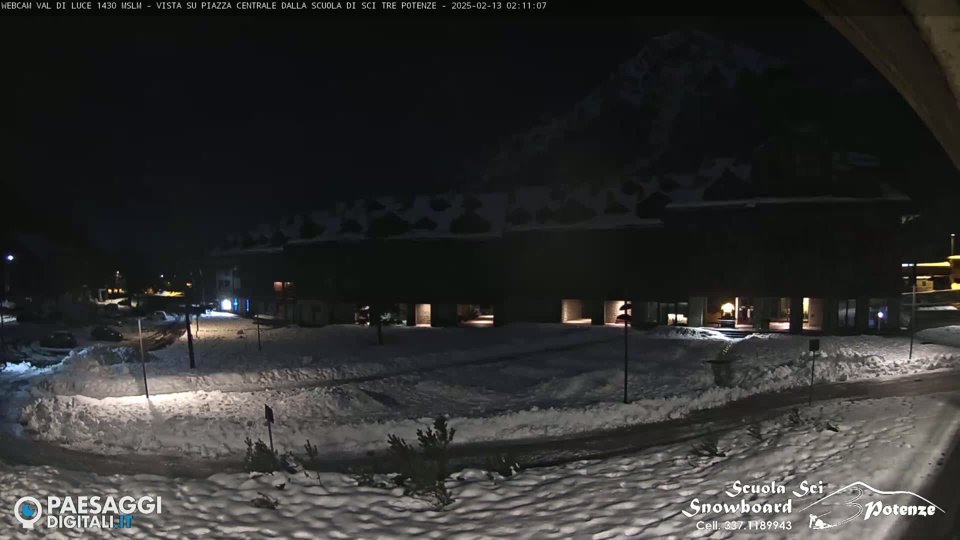 A snow-covered plaza at night, illuminated by building lights, with a dark mountain in the background.