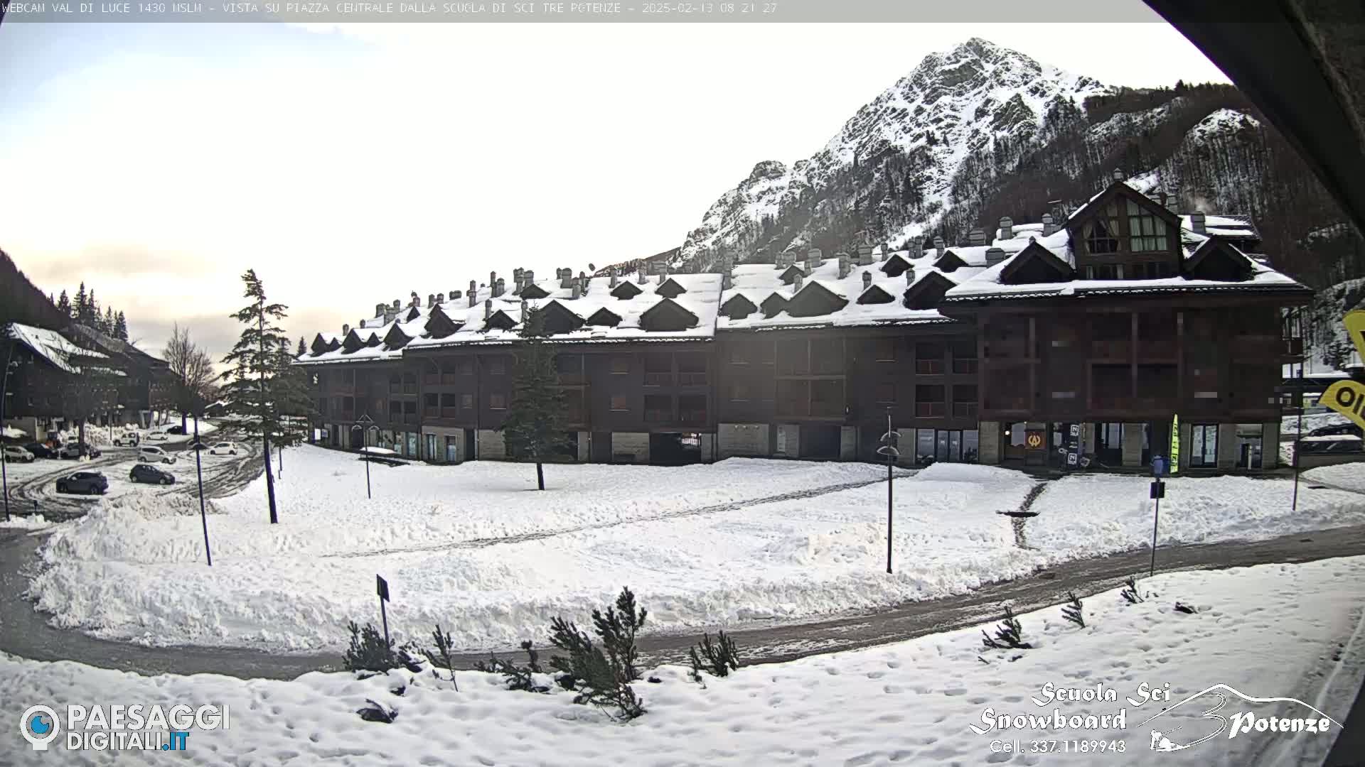A snow-covered plaza with a large apartment building and several cars parked in the foreground, mountains in the background under a cloudy sky.