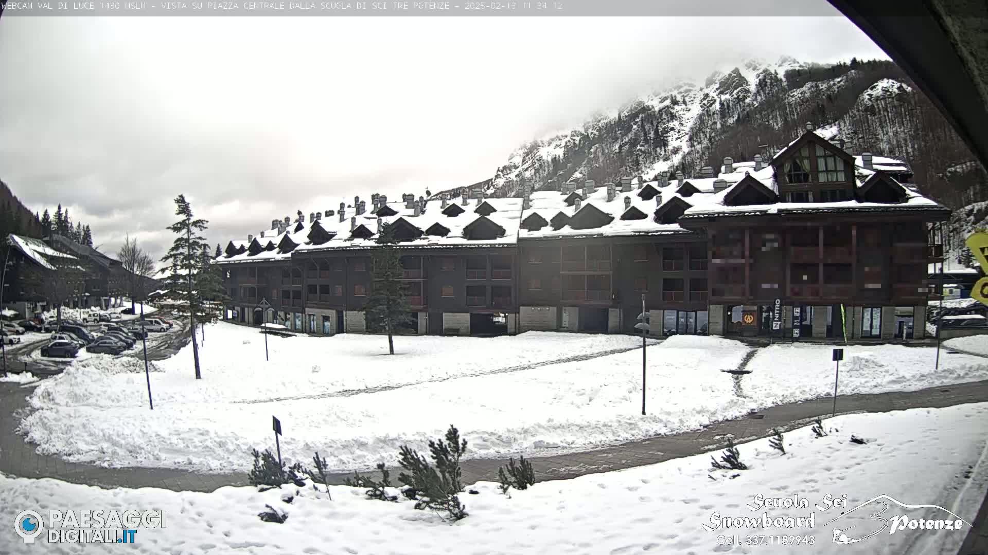 A snow-covered plaza sits in front of a long, multi-story building nestled in a mountainous area under an overcast sky.