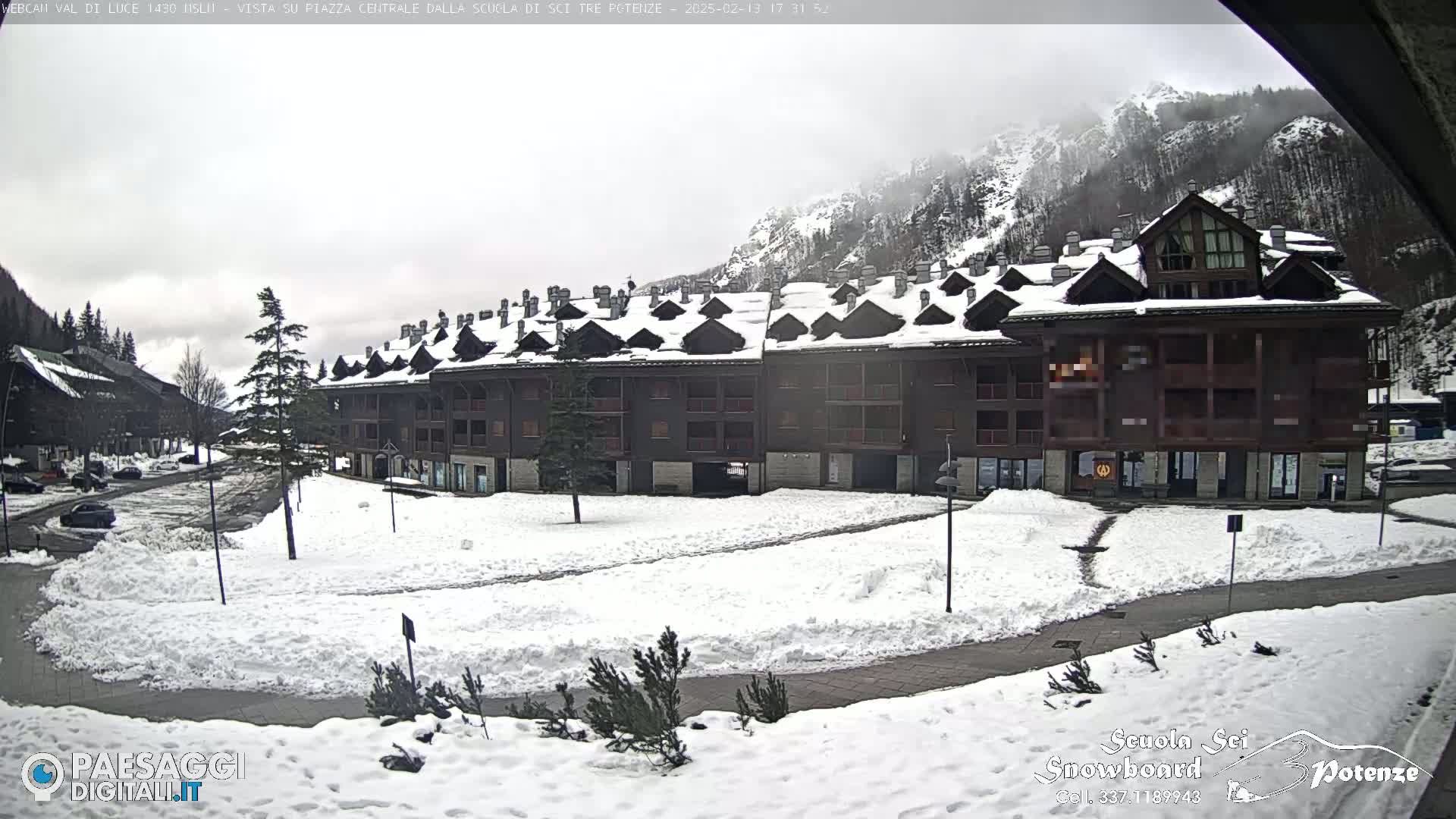 A snow-covered plaza sits in front of a multi-story building in a mountainous area under an overcast sky.