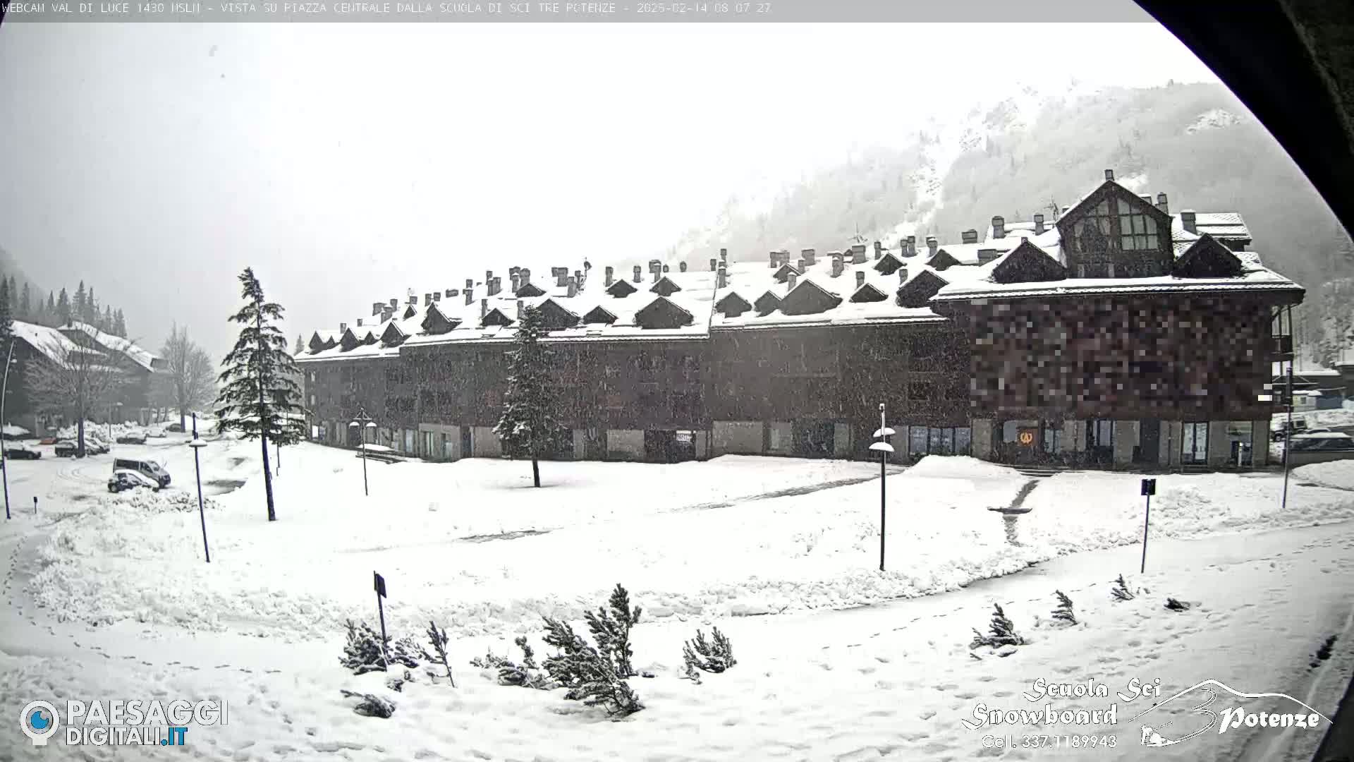 A snow-covered plaza with a large building and several cars parked, during a snowfall.