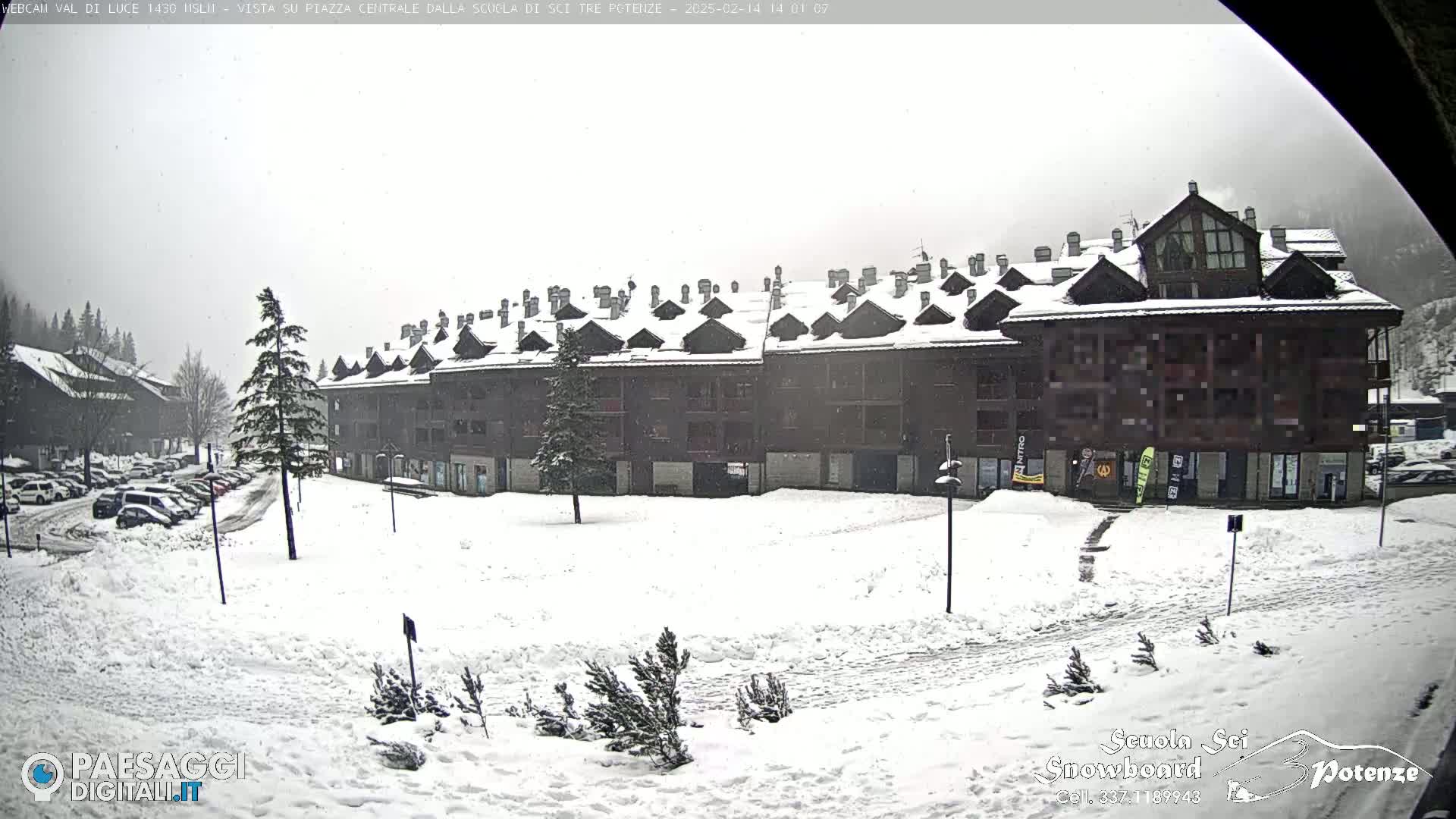 A snow-covered plaza with a multi-story building and parked cars under an overcast sky.