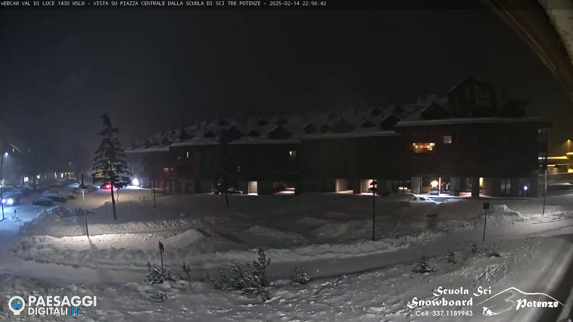A snow-covered town square at night, illuminated by streetlights, with several buildings visible and a few parked cars.