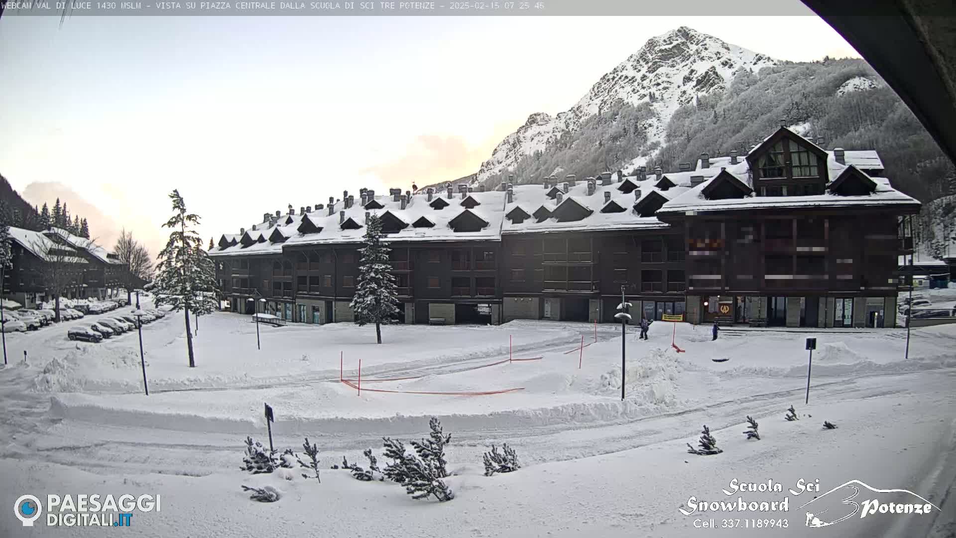 A snow-covered plaza with a large multi-story building and a mountain in the background under a partly cloudy sky.