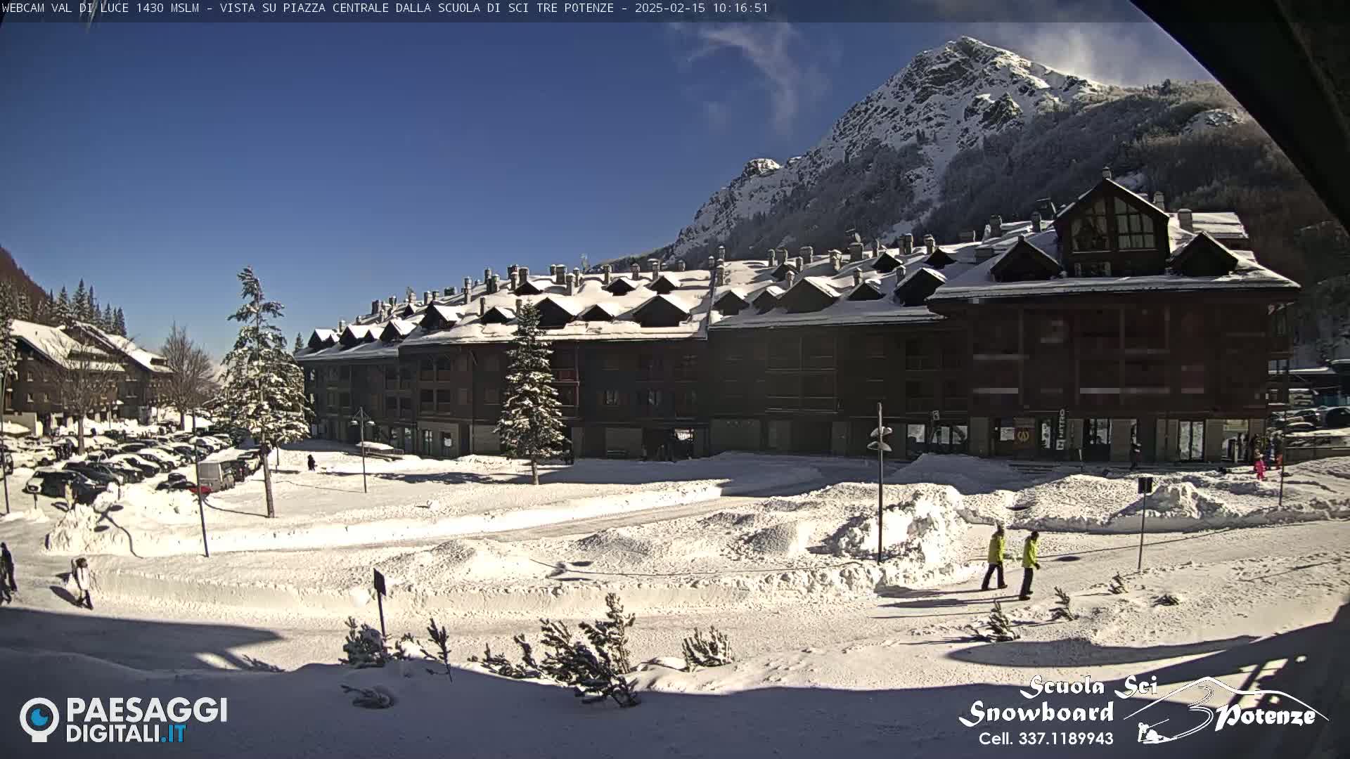 A snow-covered plaza in front of a multi-story building, with a mountain in the background under sunny skies.