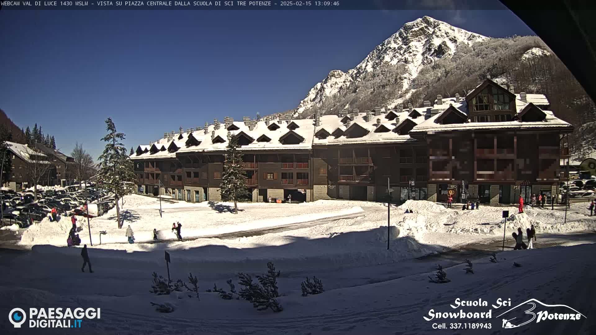 A snow-covered plaza with a multi-story building, a few people, and a mountain in the background under a sunny sky.