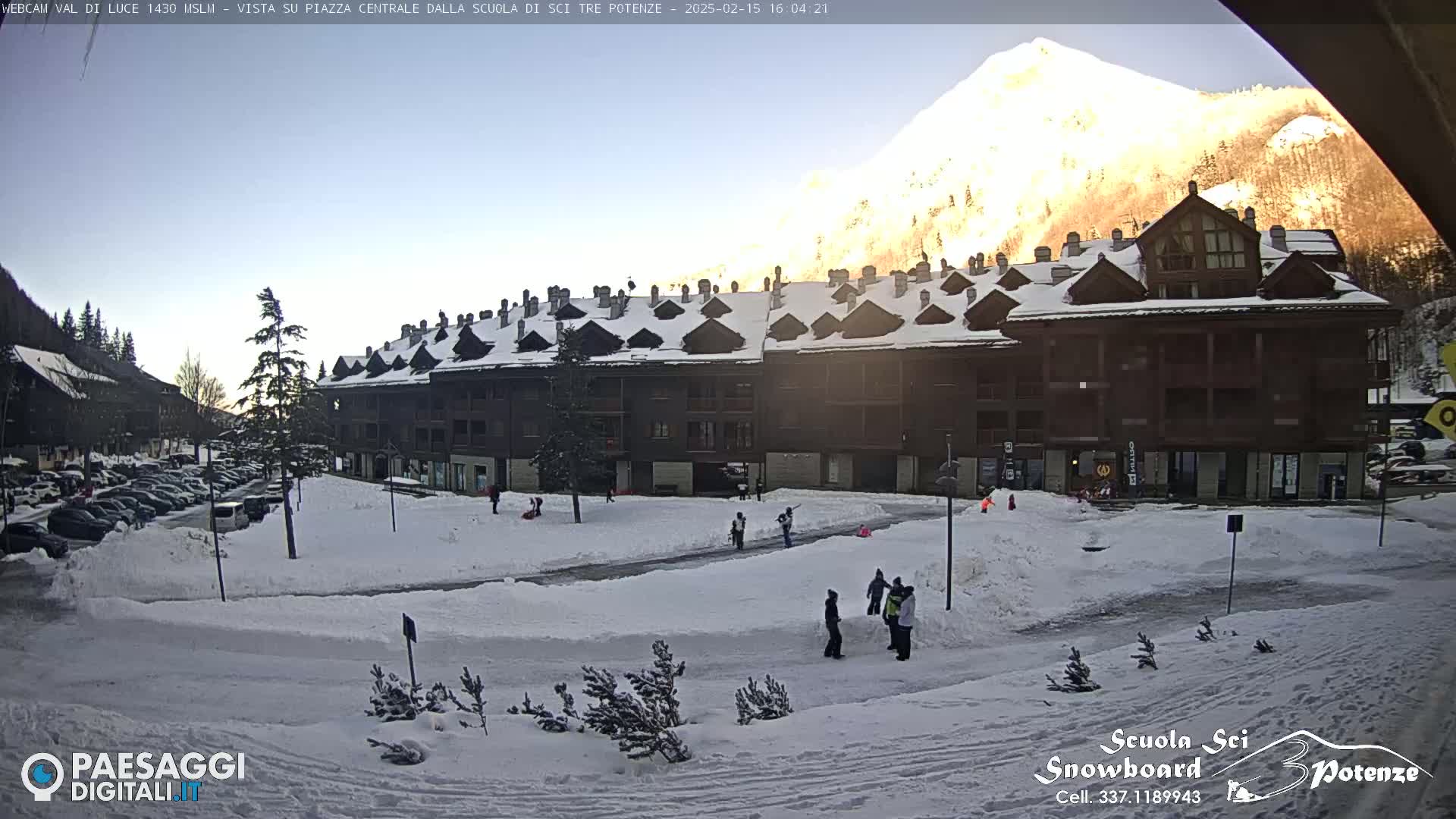 A snow-covered plaza with a multi-story building, a mountain in the background, and several people playing in the snow under a clear, sunny sky.