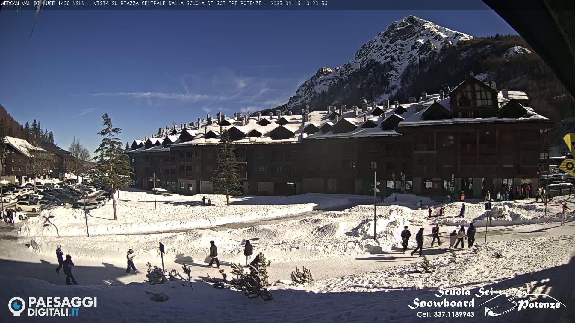 A snow-covered plaza in front of a multi-story building, with people walking around and mountains in the background under a sunny sky.