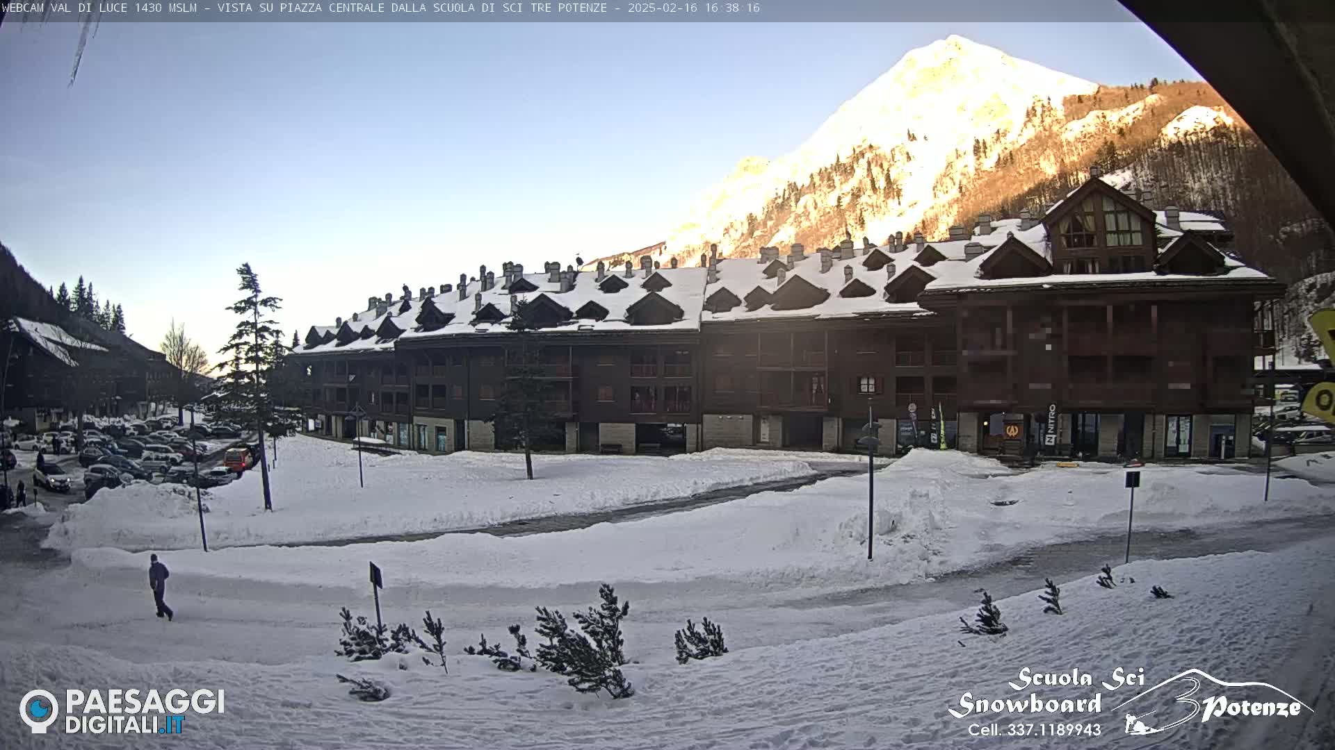 A snow-covered plaza with a large, multi-story building sits before a sunlit mountain under a clear sky.
