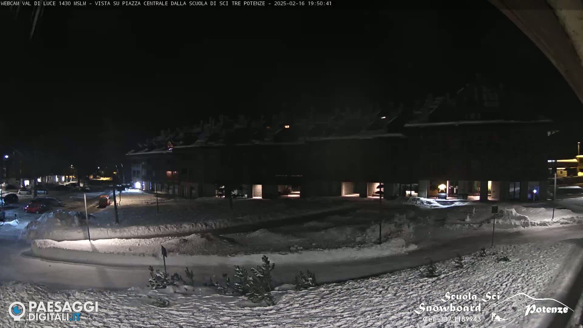 A snow-covered plaza at night, illuminated by building lights, with cars parked nearby.