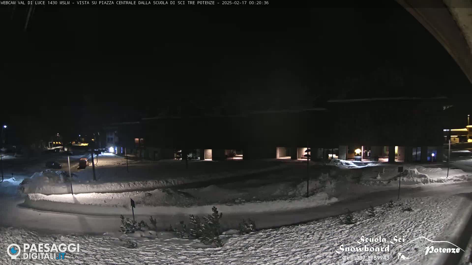 A snow-covered plaza at night, illuminated by building lights, with a few parked cars visible in the distance.