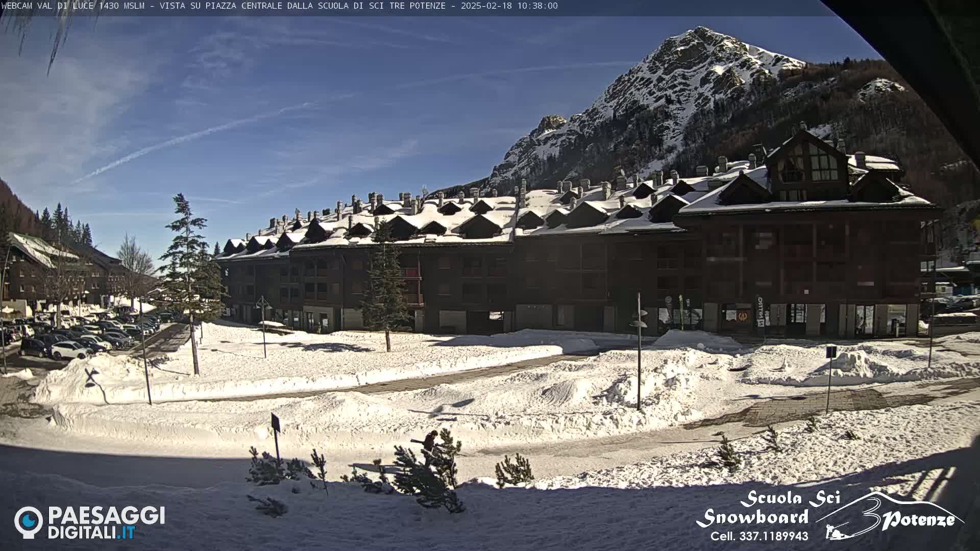 A snow-covered plaza with a large multi-story building and a mountain in the background under a sunny sky.