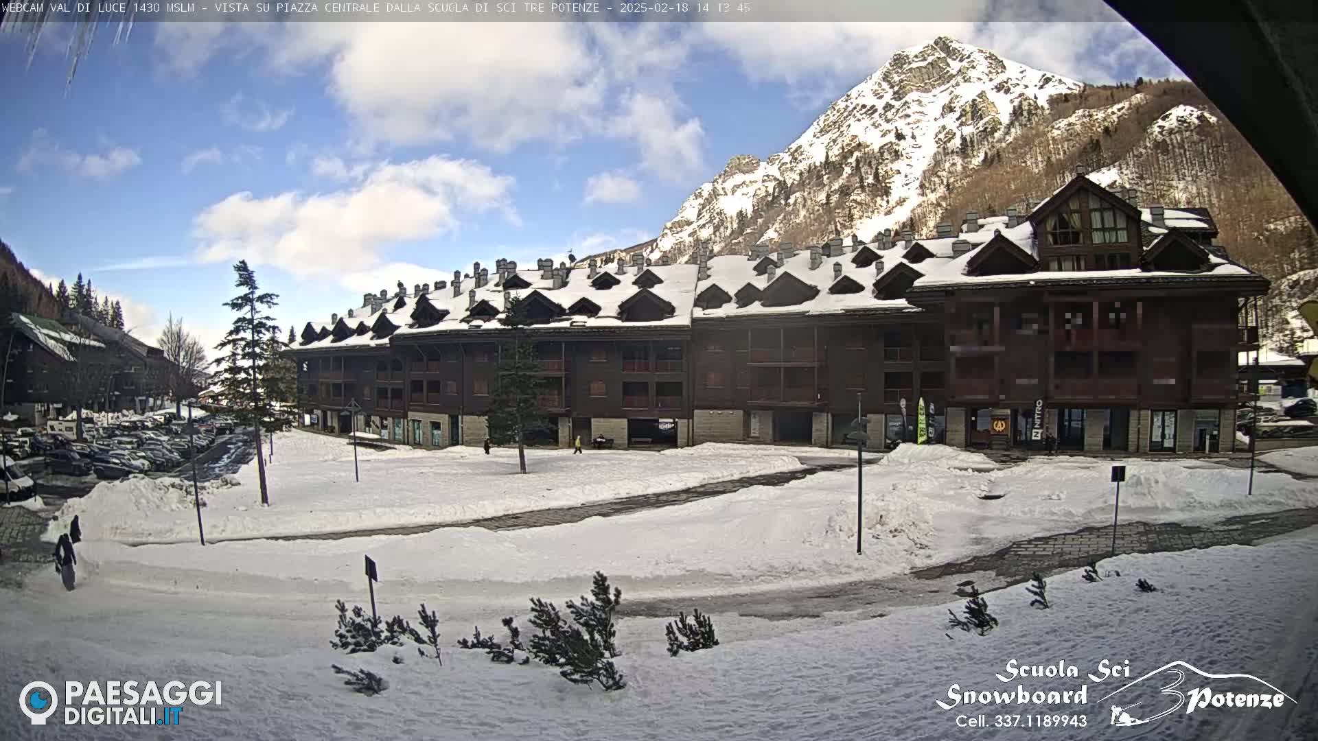 A snow-covered plaza sits in front of a multi-story brown building, with a snow-capped mountain in the background under a partly cloudy sky.