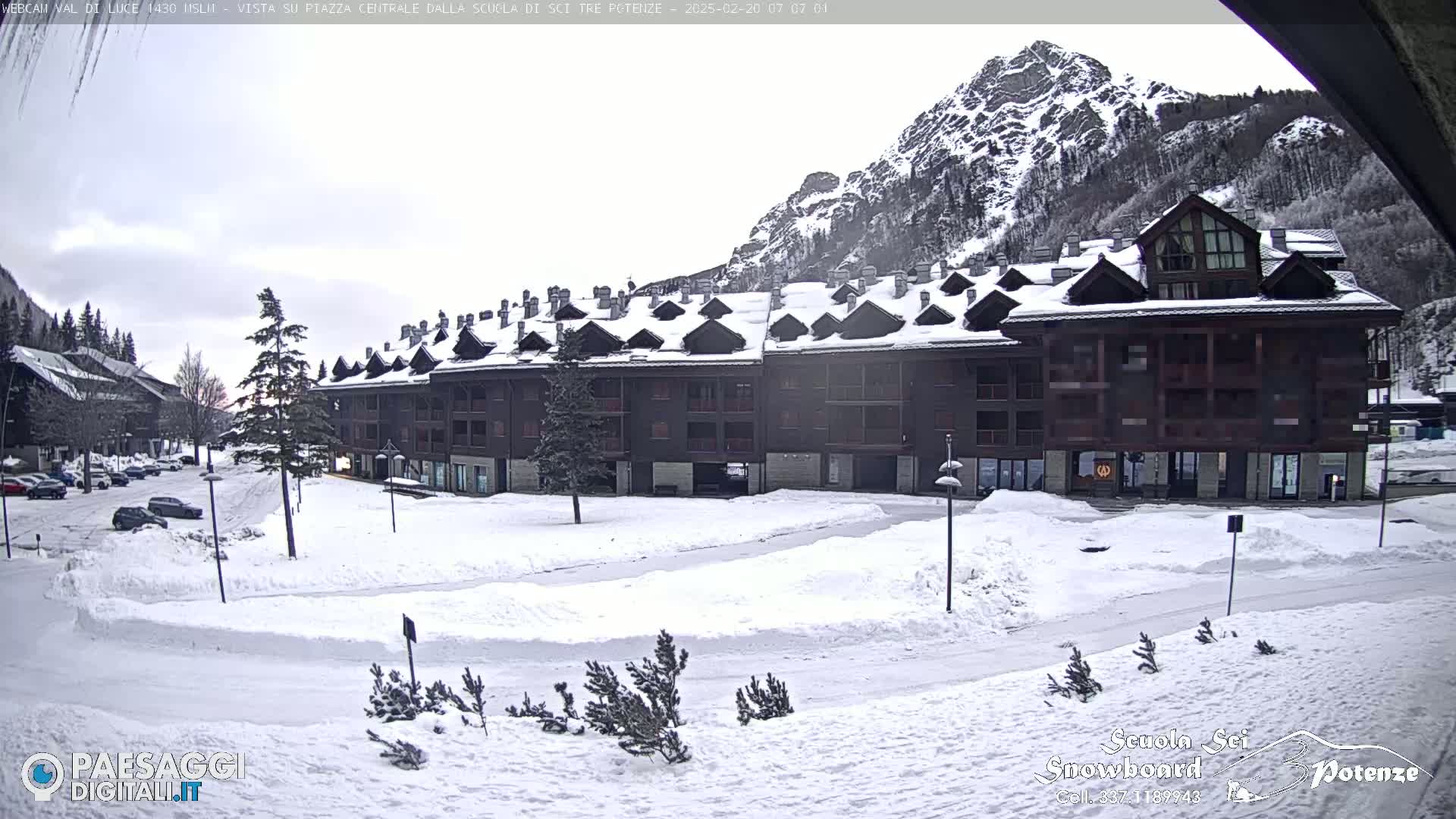 A snow-covered plaza with a large multi-story building and mountains in the background under a cloudy sky.