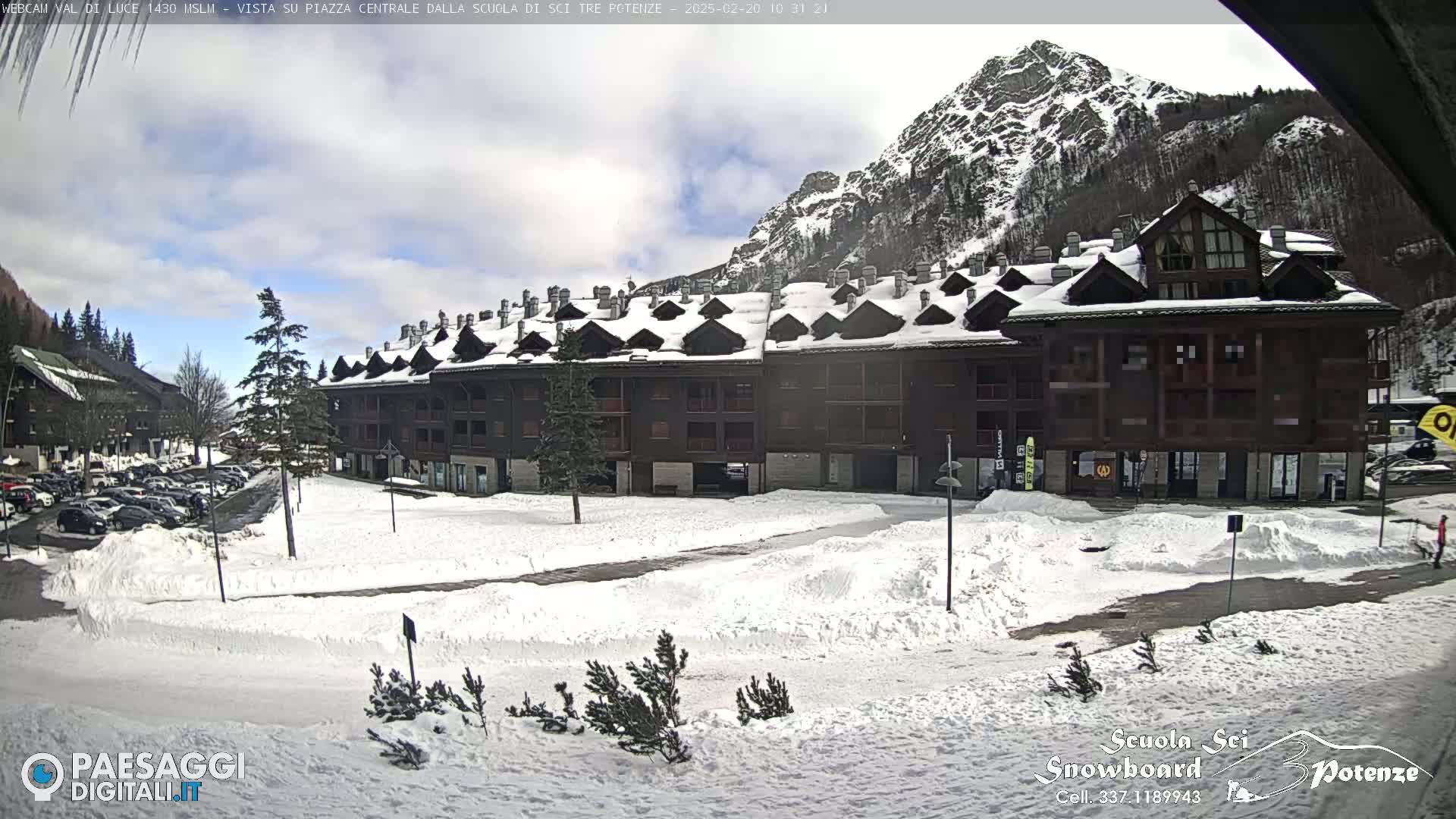 A snow-covered plaza with a multi-story building and a mountain in the background under a partly cloudy sky.