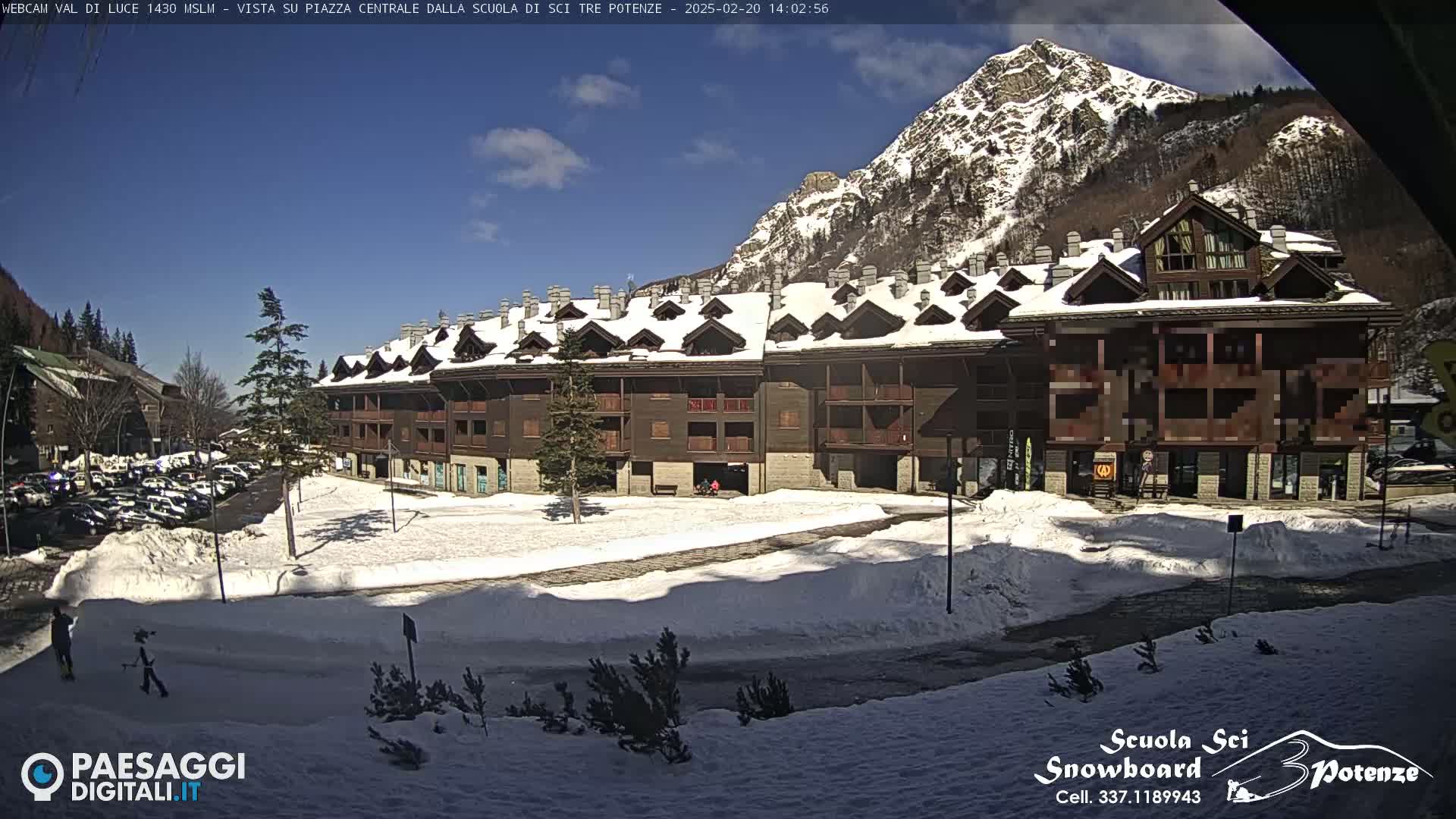 A snow-covered plaza with a multi-story building is in front of a sunny, snow-capped mountain.