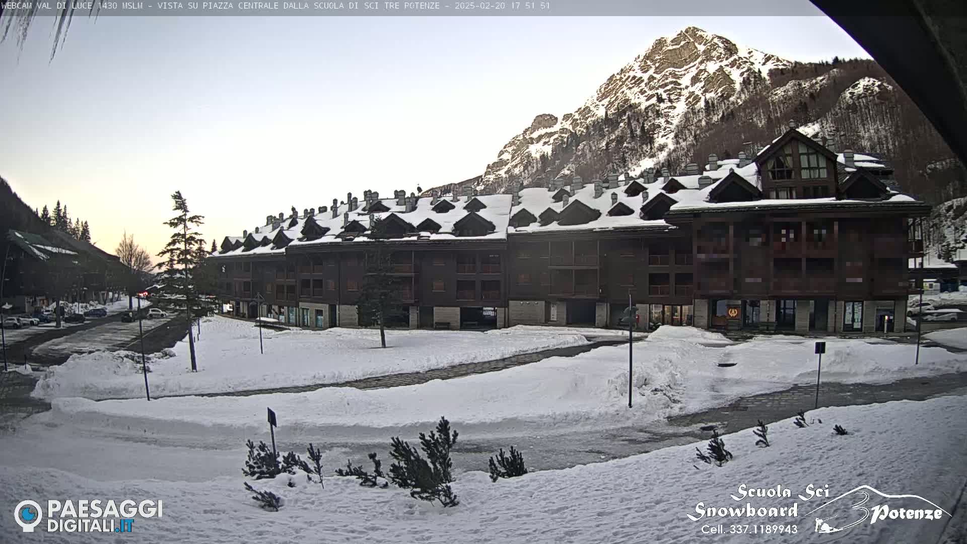 A snow-covered plaza sits in front of a multi-story brown building, with a mountain in the background under a mostly clear sky.
