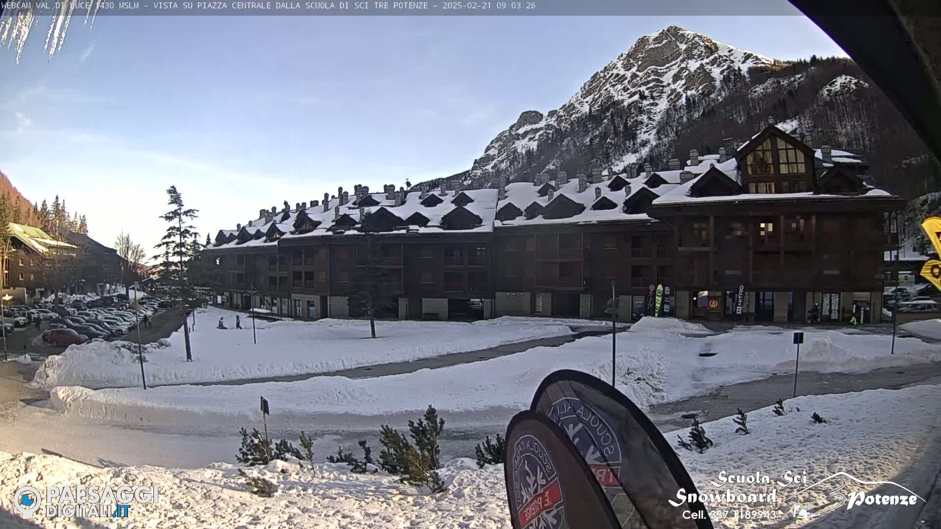 A snow-covered plaza is situated in front of a multi-story building, with a mountain in the background under a mostly clear sky.