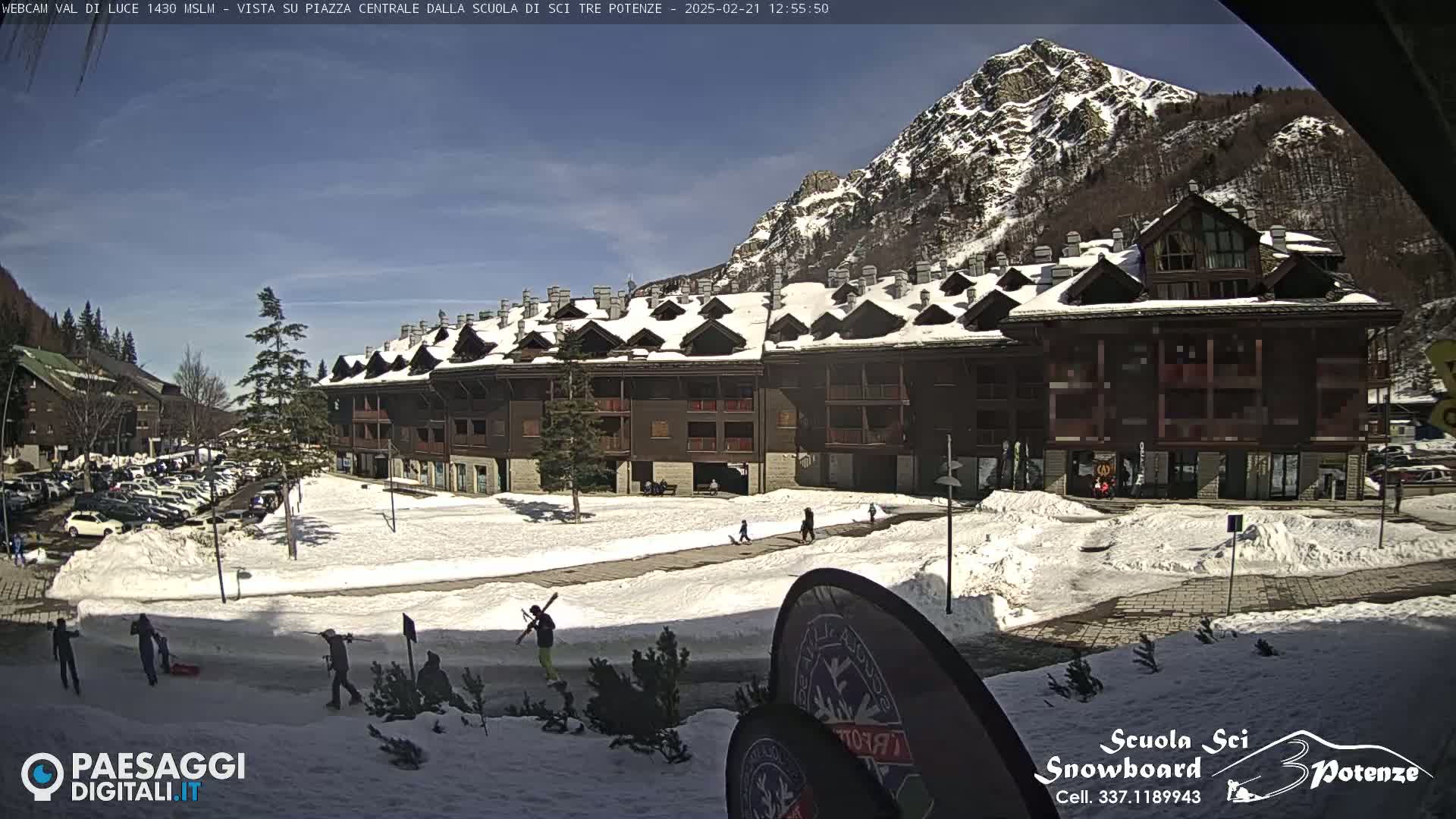 A snow-covered plaza in front of a multi-story brown building, with a mountain in the background under a sunny sky.