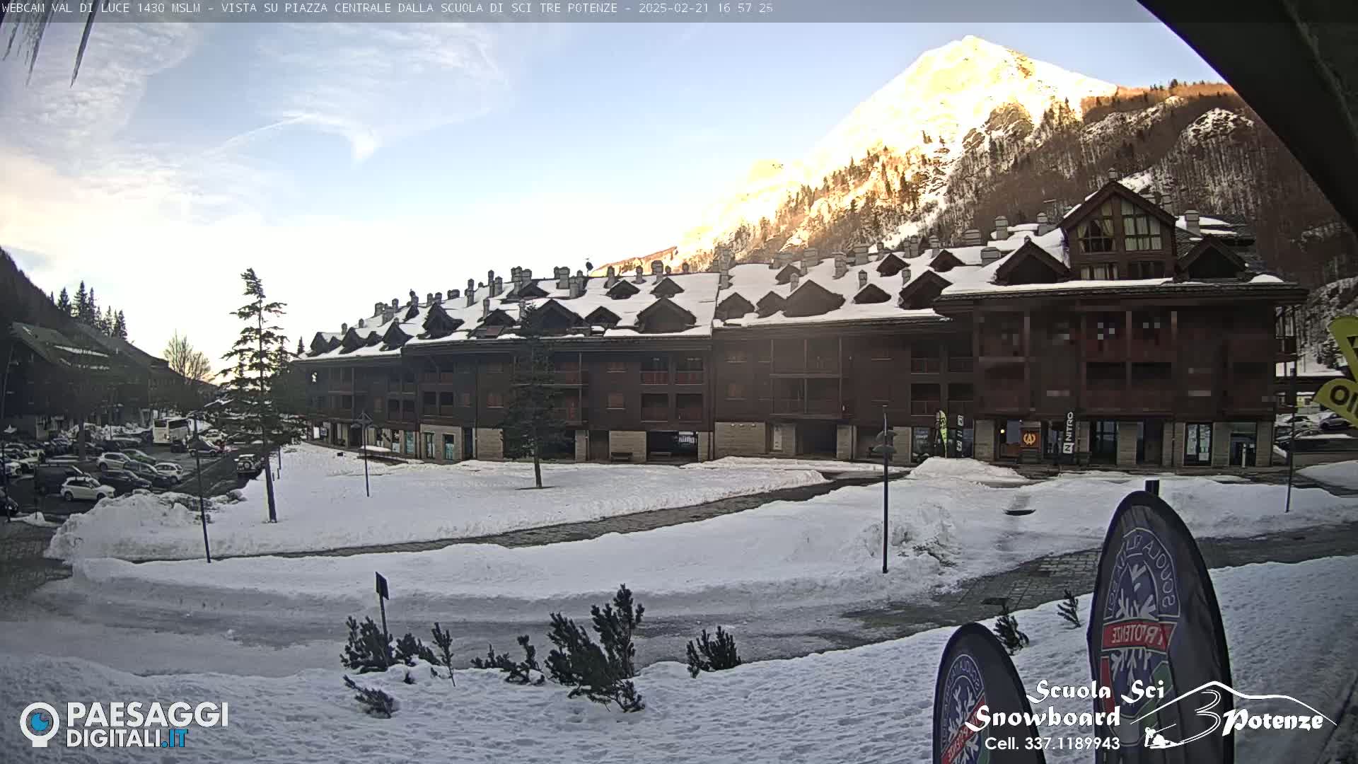 A snow-covered plaza sits in front of a multi-story building in a mountainous area under a partly sunny sky.