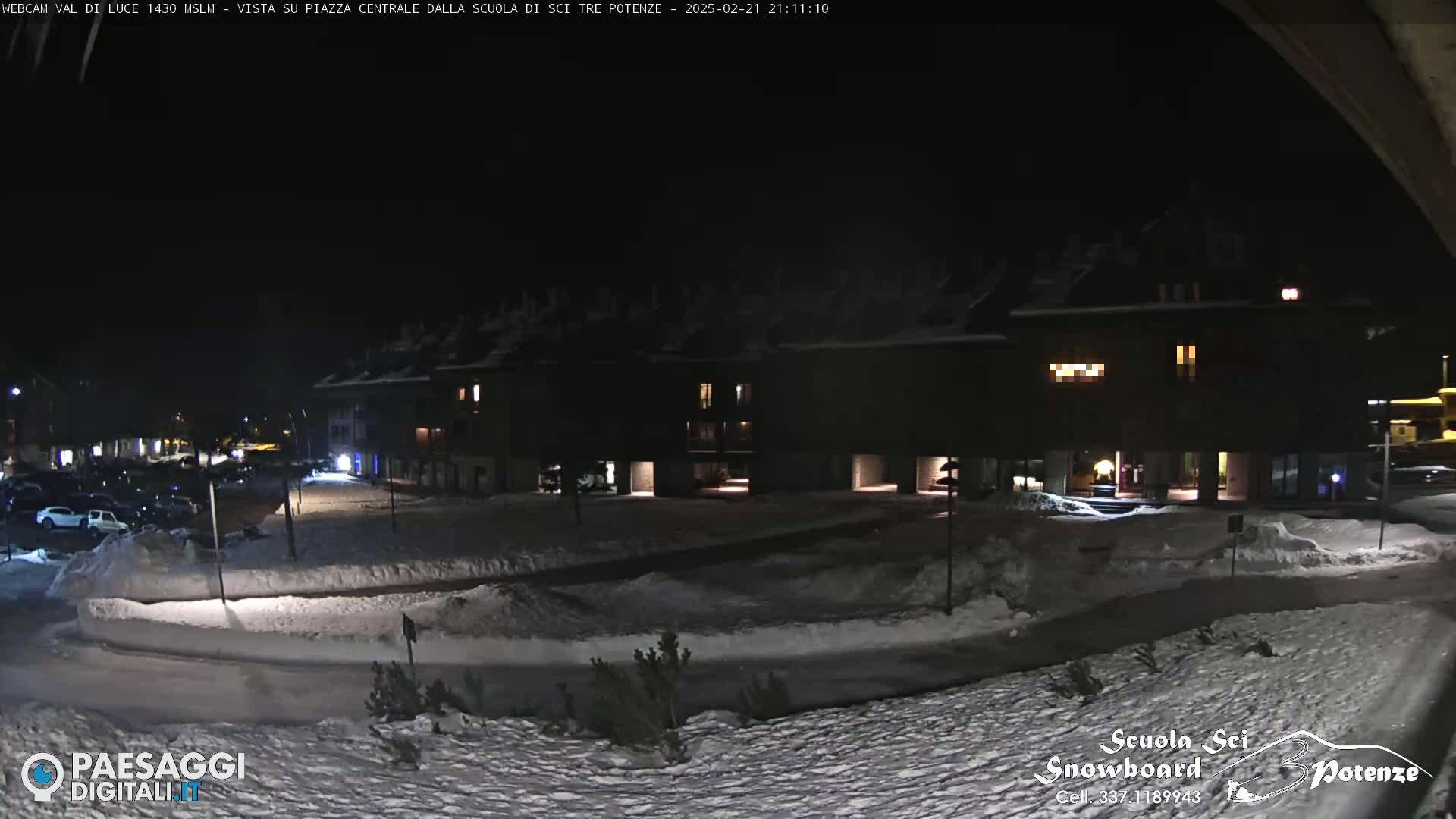 A snow-covered nighttime scene shows a parking lot and several buildings with some lights on.