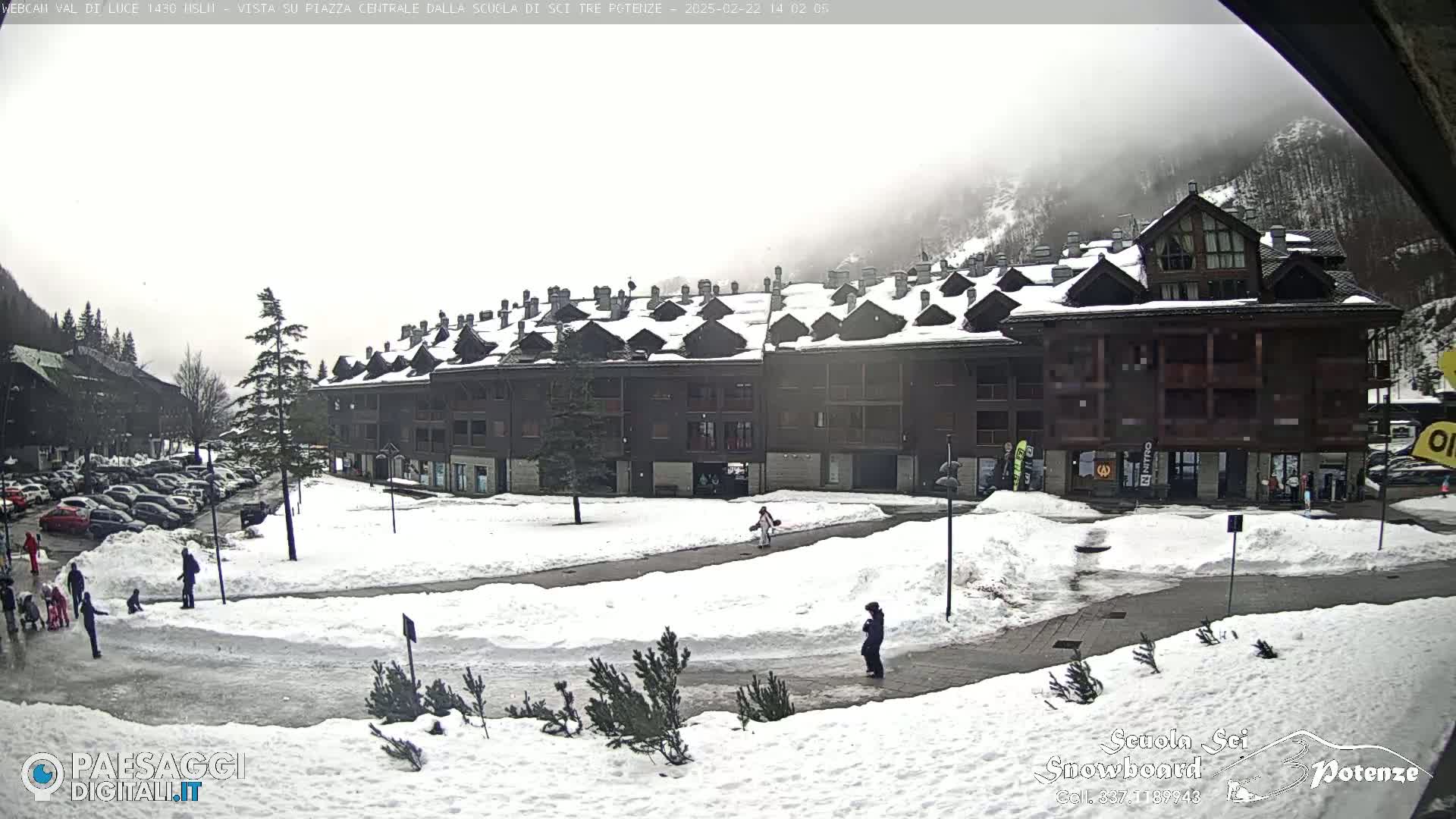 A snow-covered plaza in front of a multi-story building, with a few people walking around under an overcast sky.
