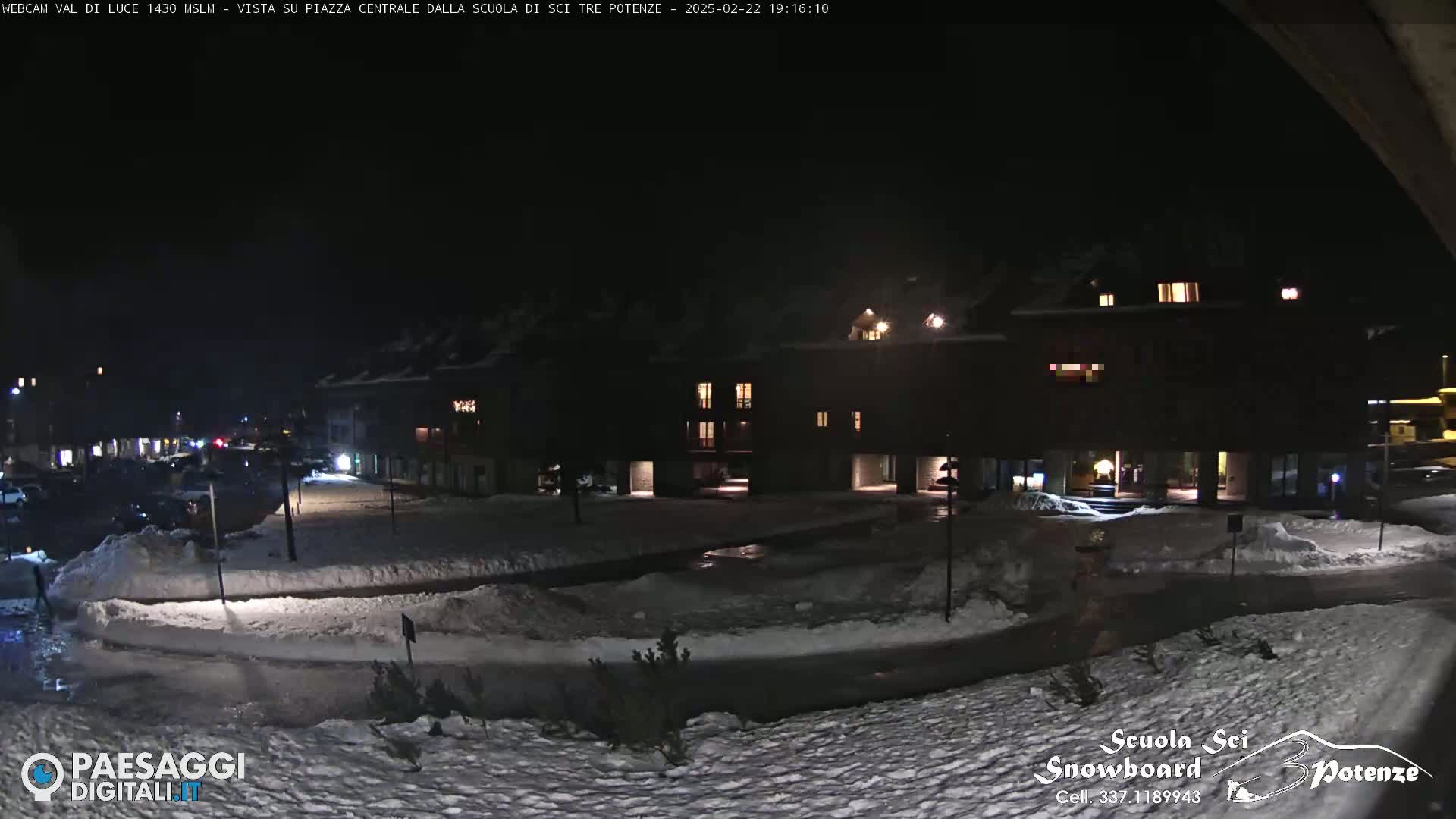 A snow-covered town square at night, illuminated by building lights, with some cars parked nearby.