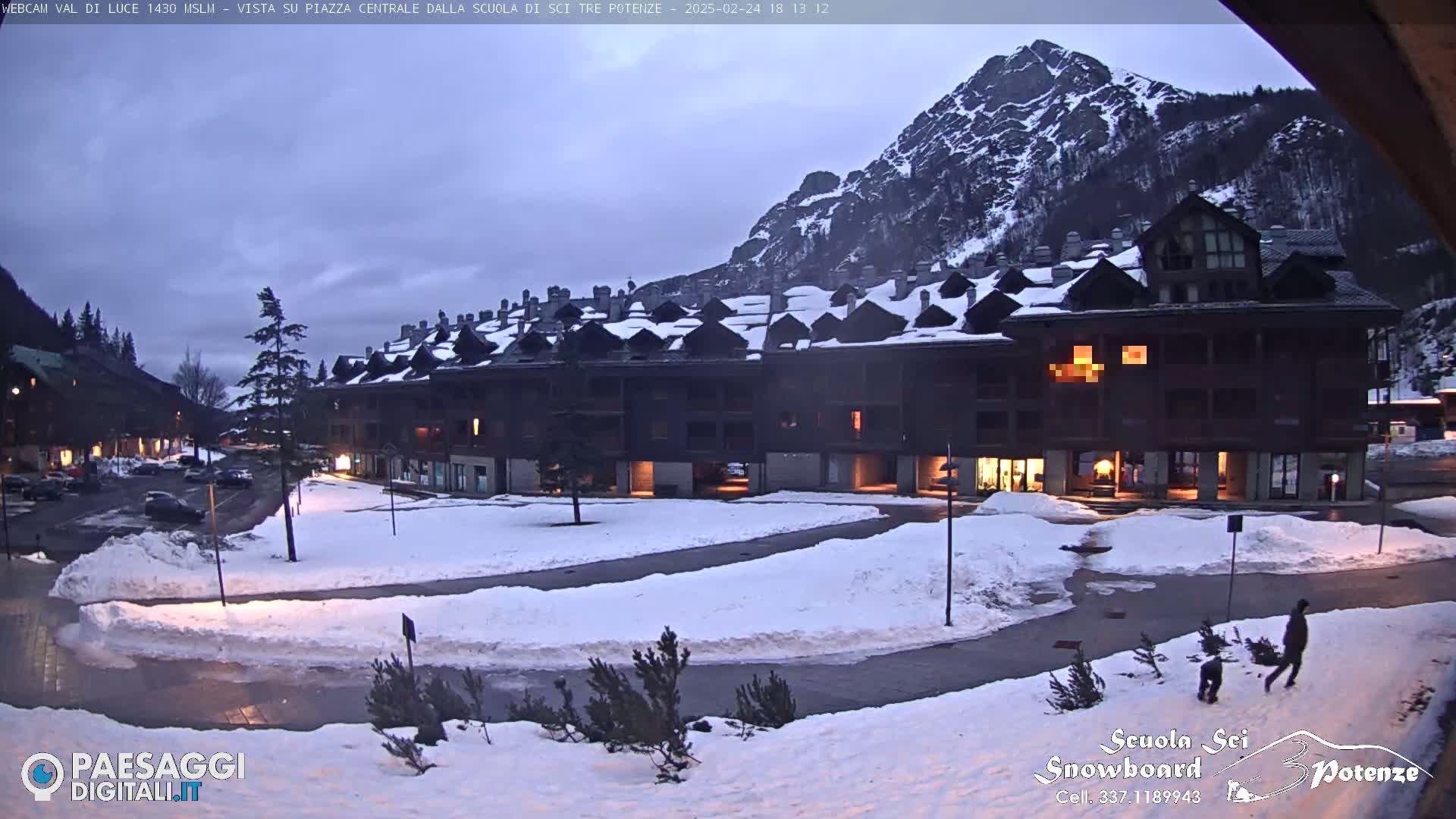 A snow-covered plaza in front of a multi-story building at dusk, with a mountain in the background under a cloudy sky.