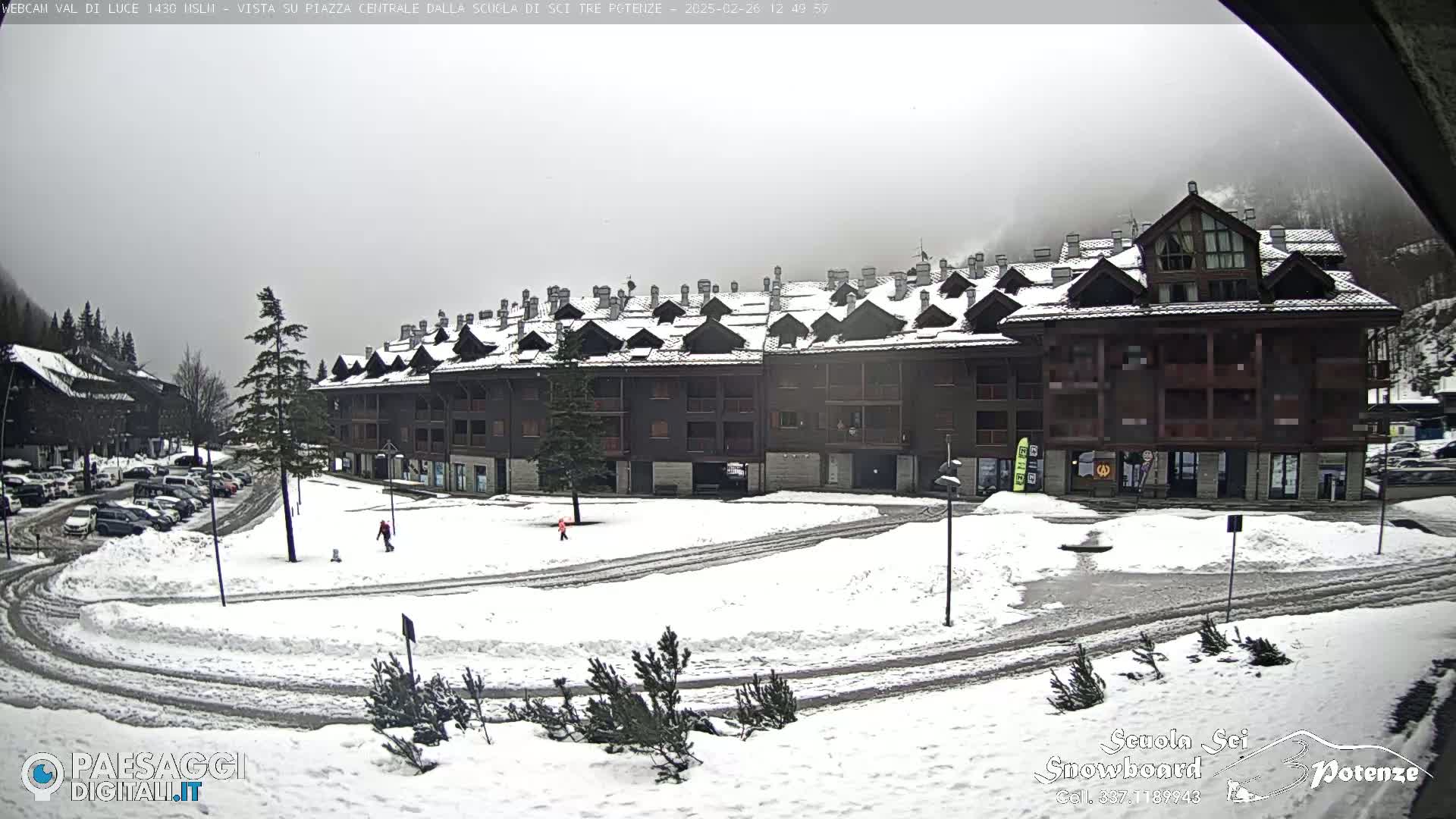 A snow-covered parking lot and plaza are in front of a large, multi-unit building under an overcast sky.