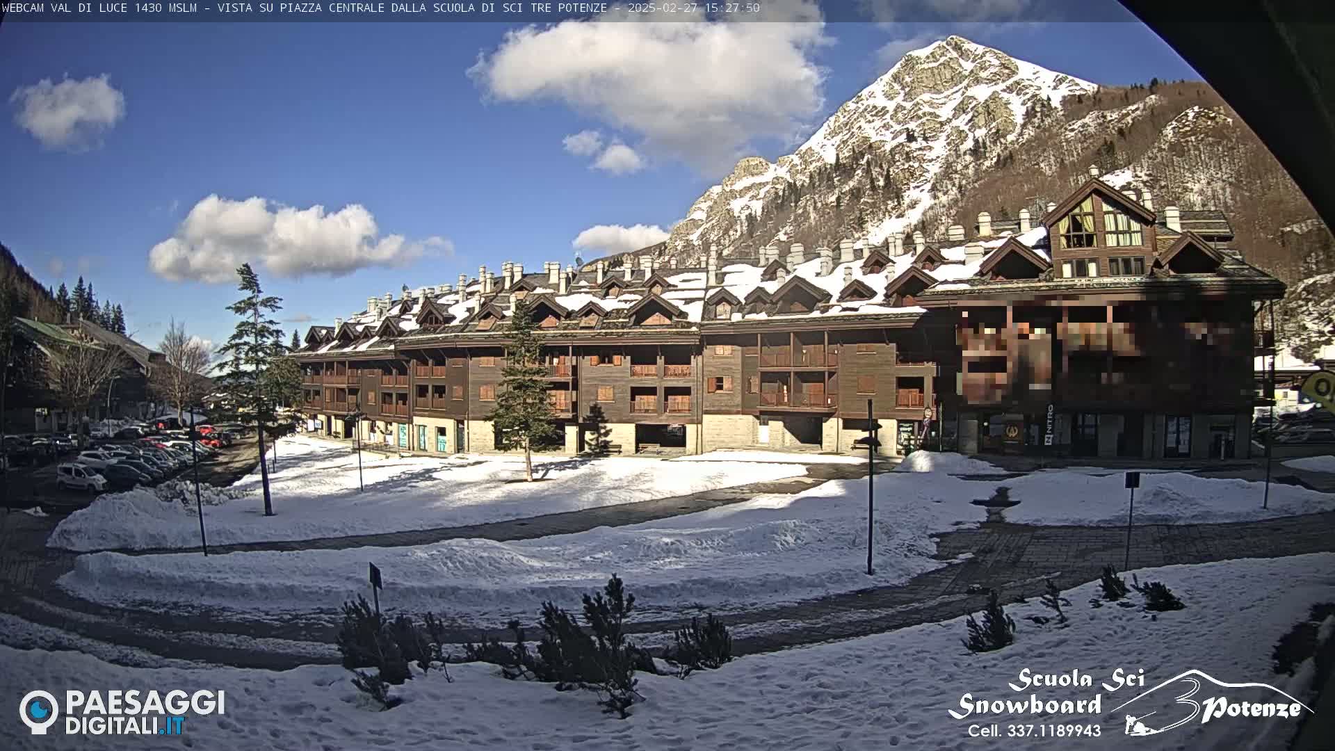A large, wood-sided building sits in a snowy square, with a mountain backdrop under a partly cloudy, sunny sky.