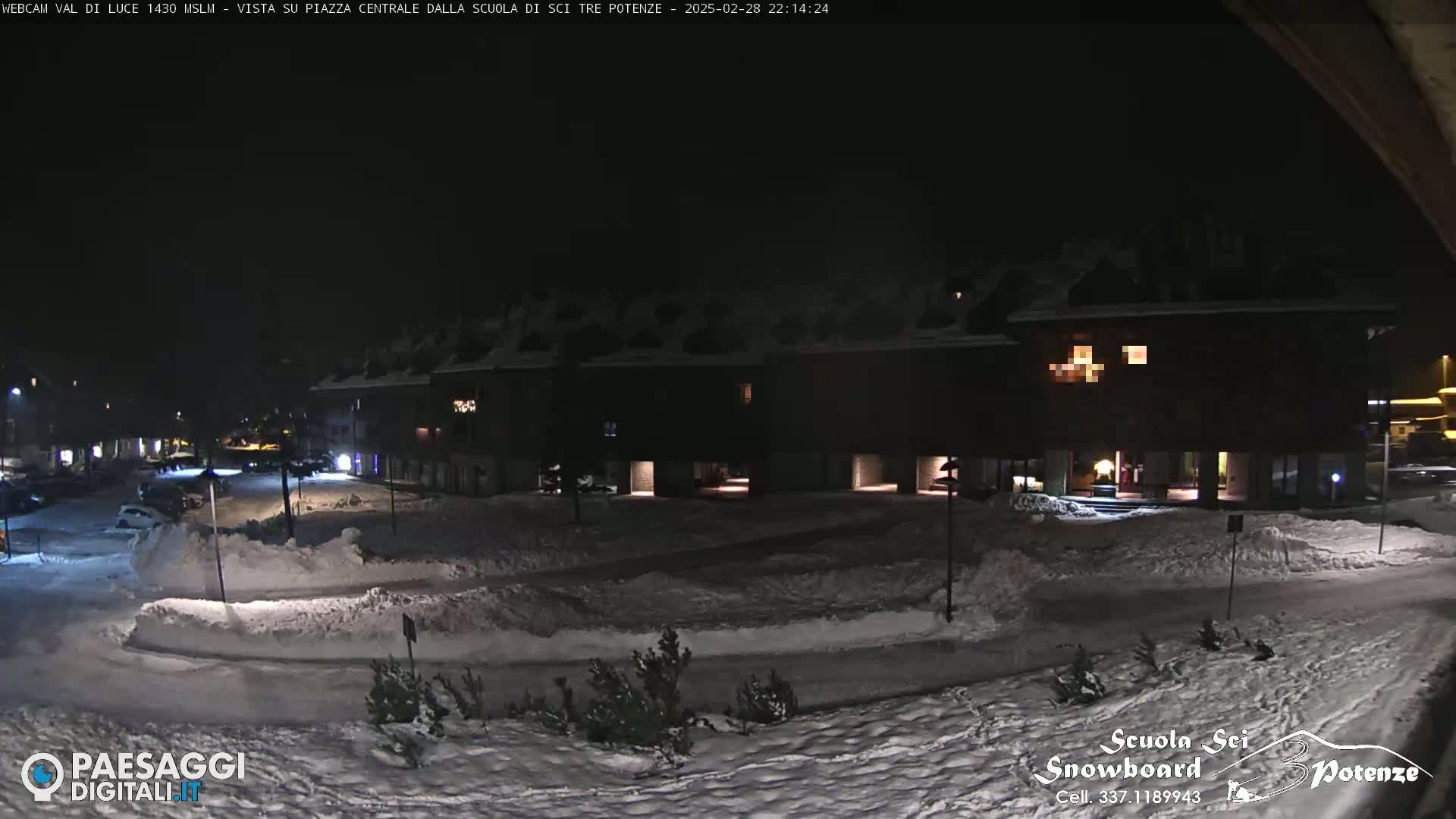 A snow-covered town square at night, illuminated by streetlights, with a large building in the background.