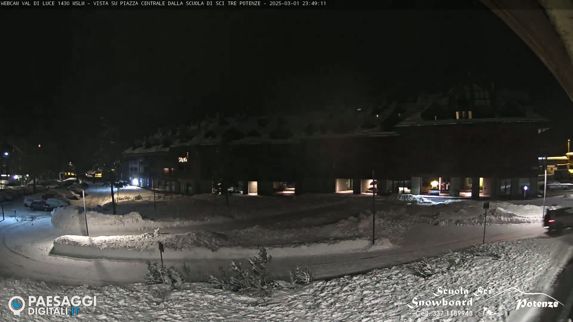 A snow-covered plaza at night is illuminated by streetlights, with a building and parked cars visible in the background.