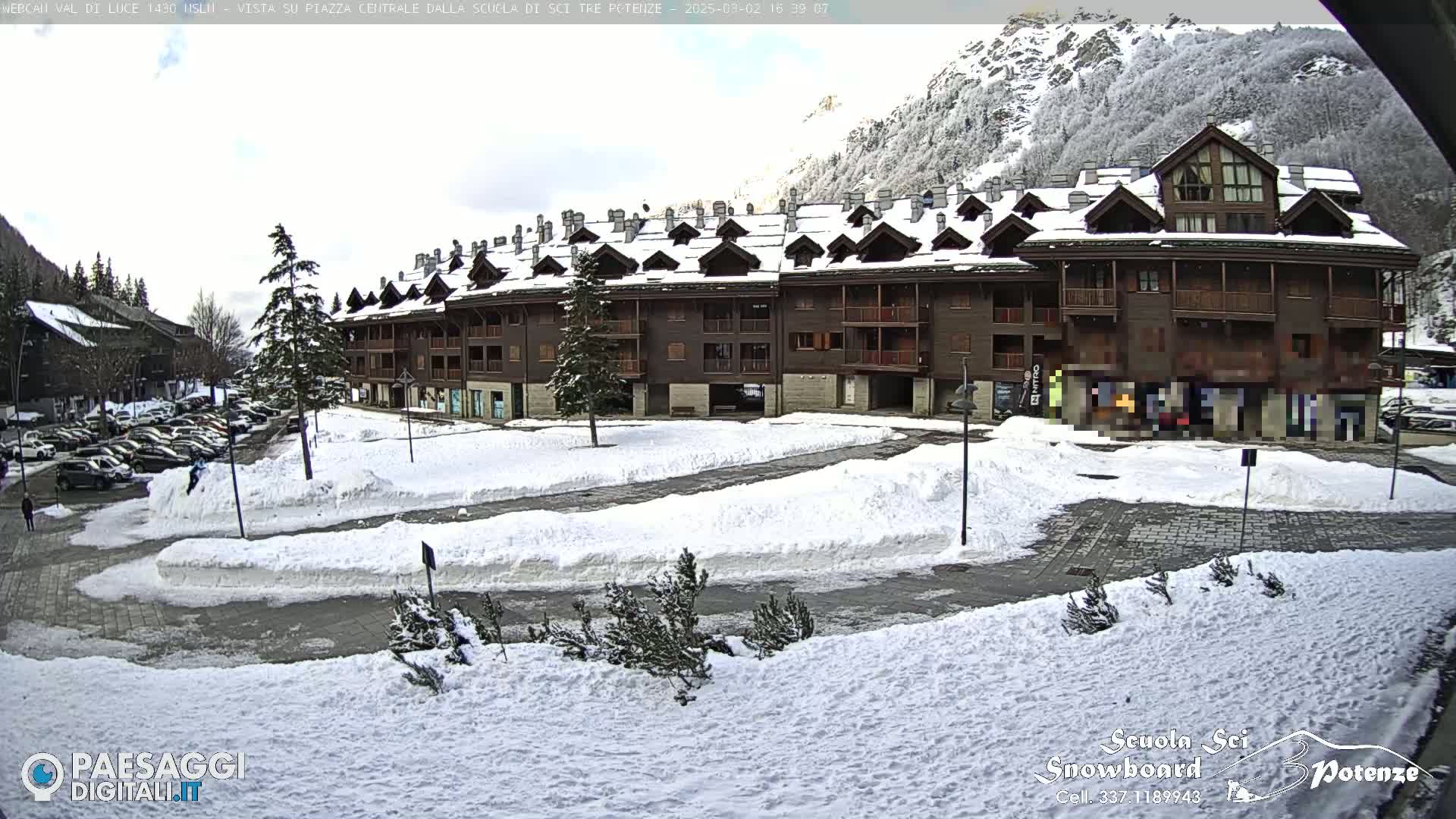 A snow-covered parking lot and plaza are in front of a multi-story brown building, with snow-covered mountains in the background.