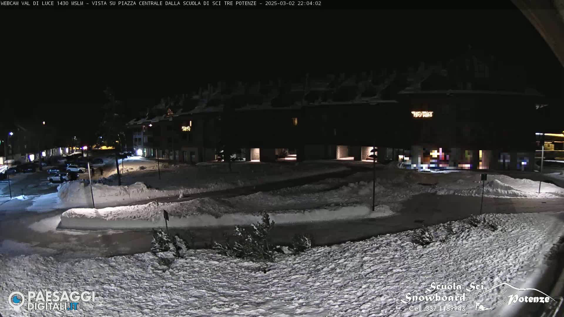 A snow-covered town square at night, illuminated by streetlights, with buildings lining the perimeter.