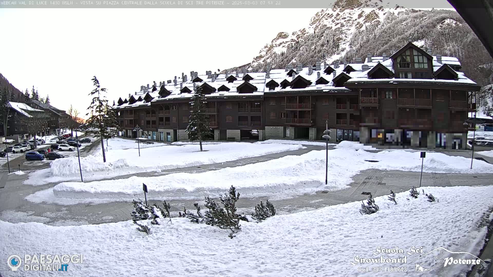 A snow-covered parking lot and plaza are in front of a multi-story brown building, set against a backdrop of snow-covered mountains.