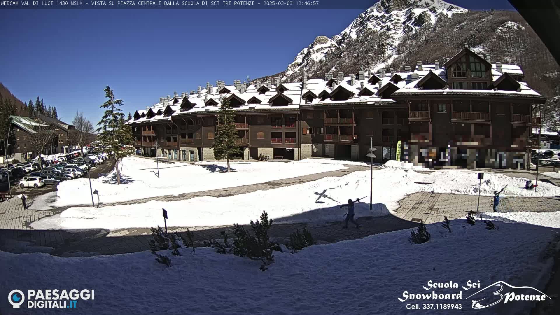 A snow-covered plaza in front of a multi-story building, with a sunny sky and mountains in the background.