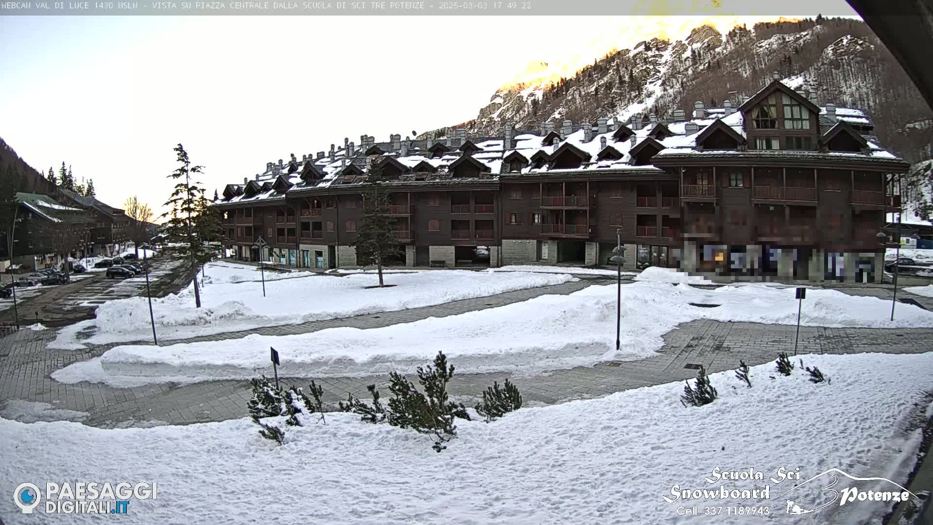 A snow-covered plaza sits in front of a large, multi-story, brown wood building nestled in a mountain valley at sunset.