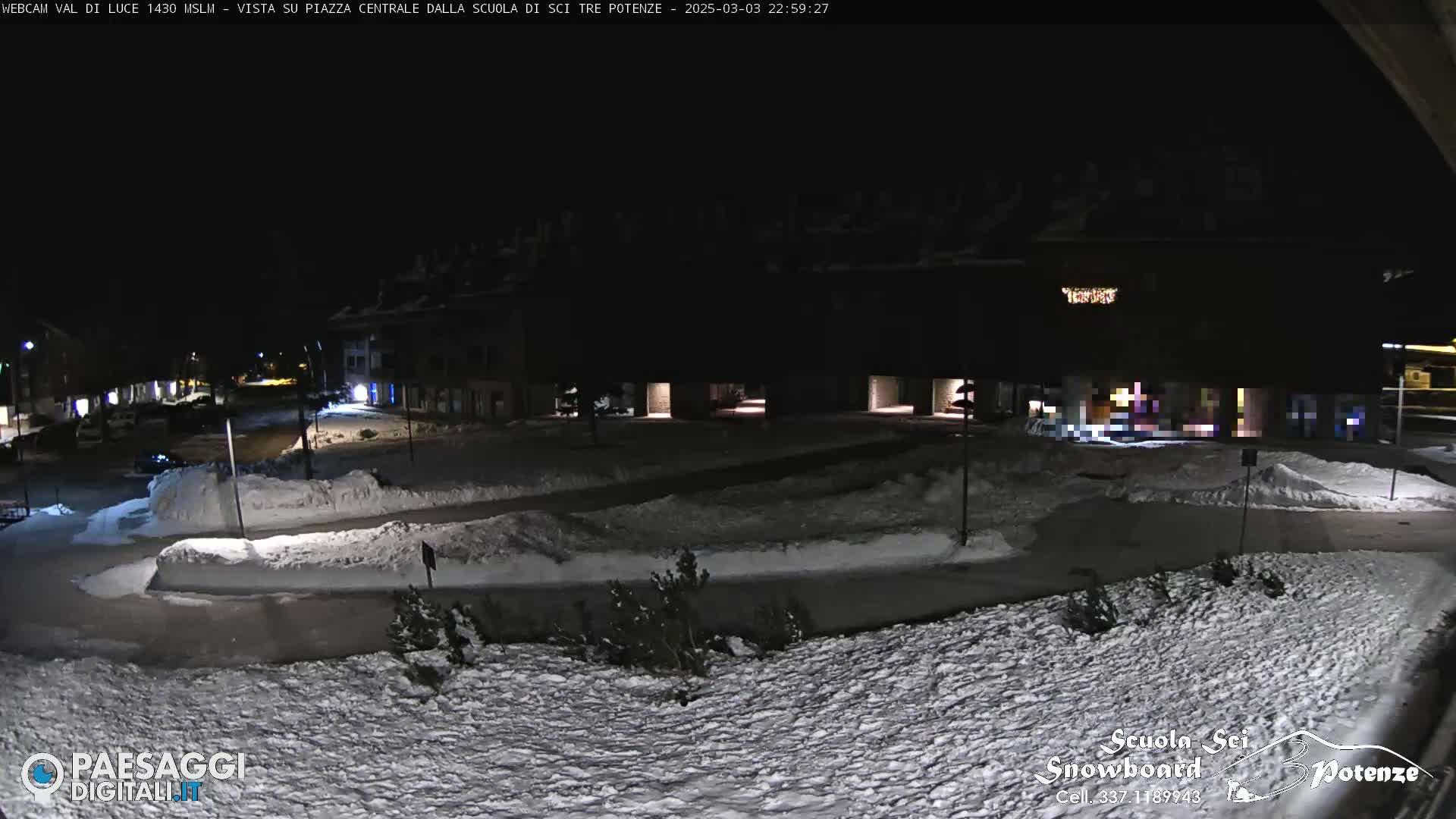 A snow-covered plaza at night, illuminated by streetlights, with buildings visible in the background.