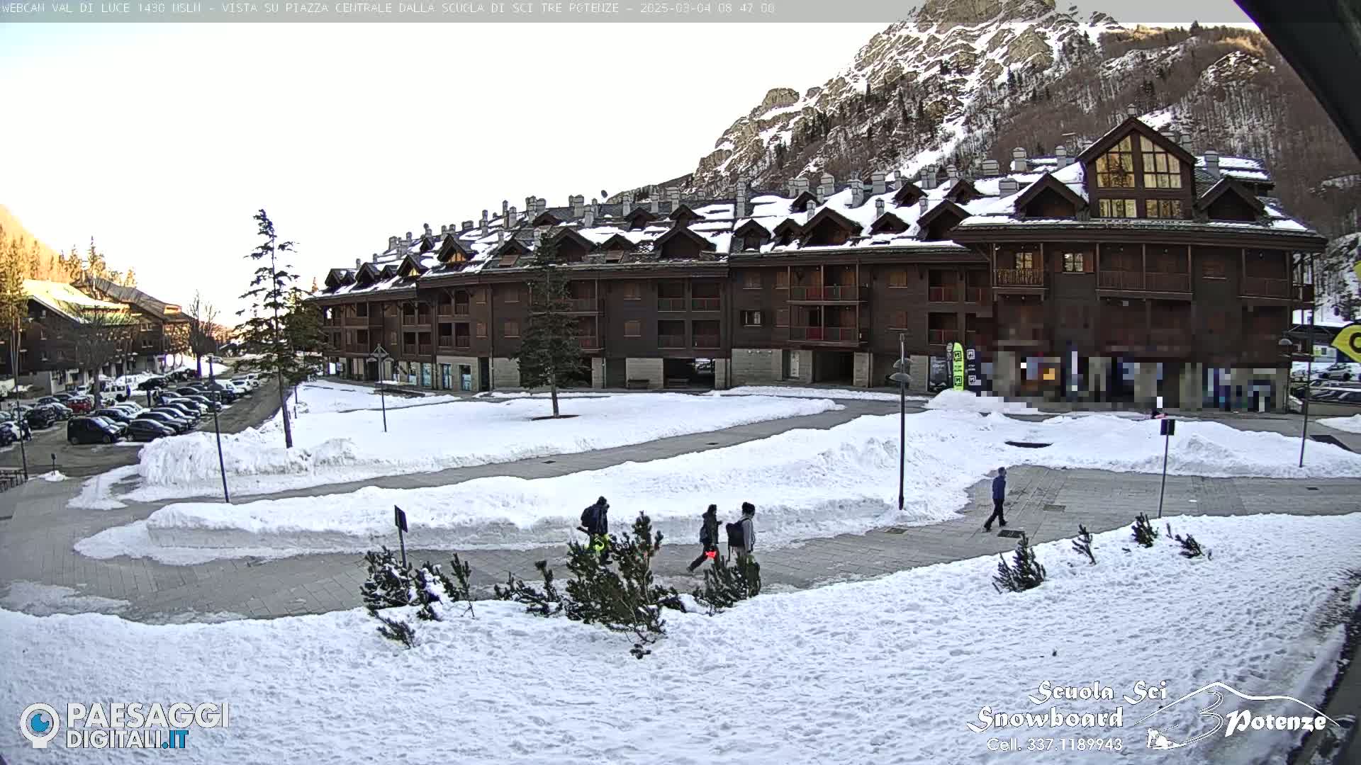 A snow-covered plaza with a multi-story building and several people walking, situated in front of a mountain range under a mostly clear sky.