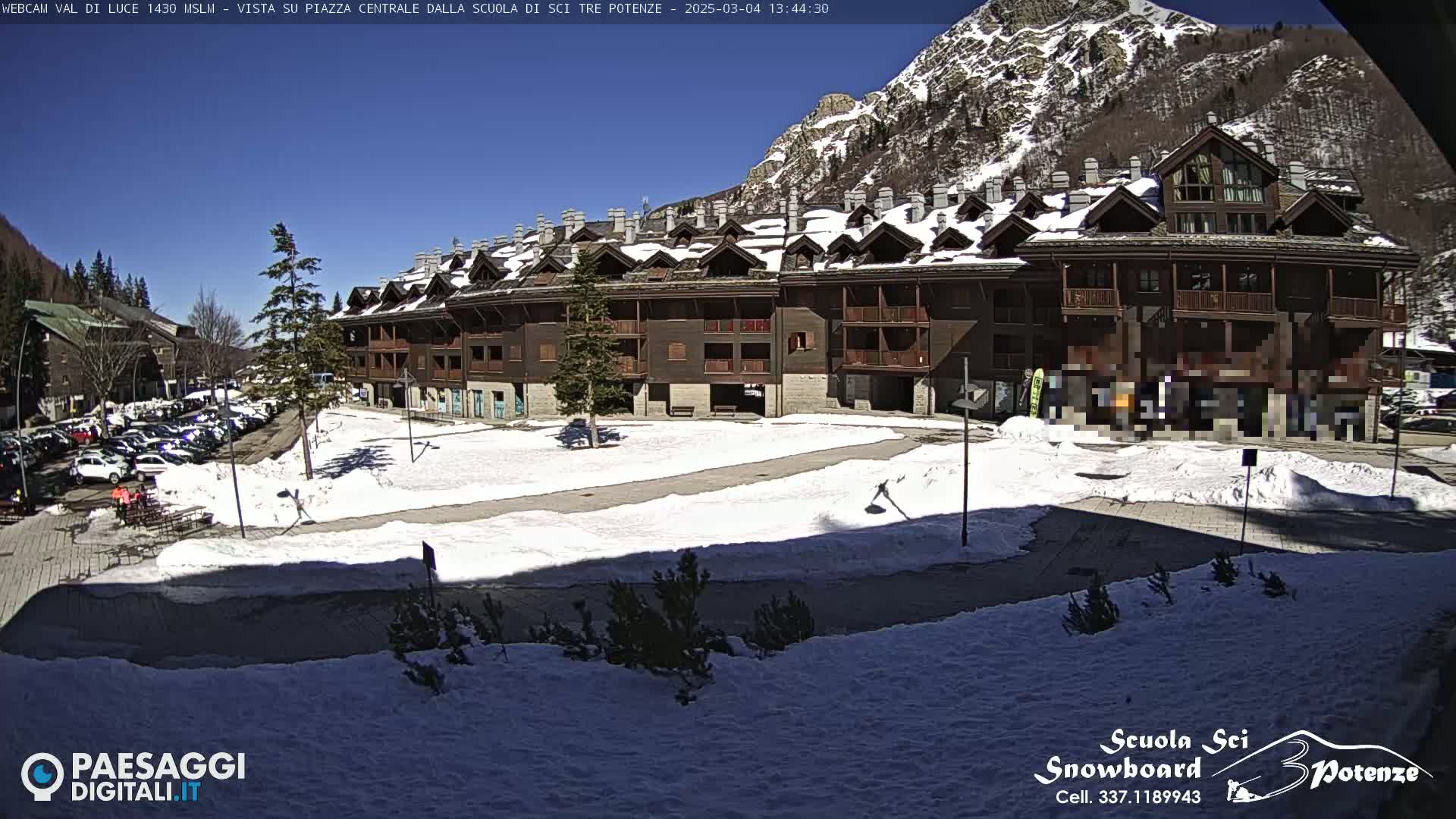 A large, multi-story brown building is situated in a snowy plaza, with a mountain backdrop under a clear, sunny sky.