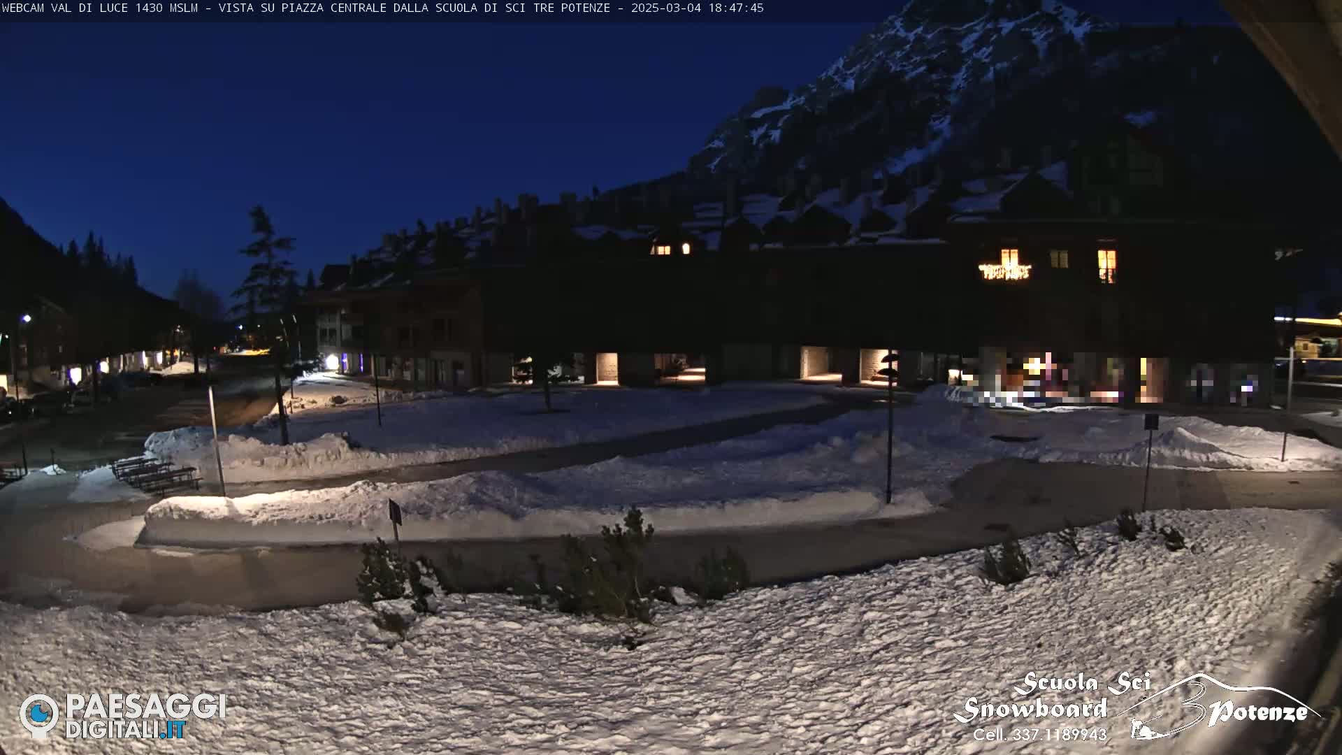 A snow-covered town square at night, illuminated by a few building lights, is nestled beneath a dark mountain.