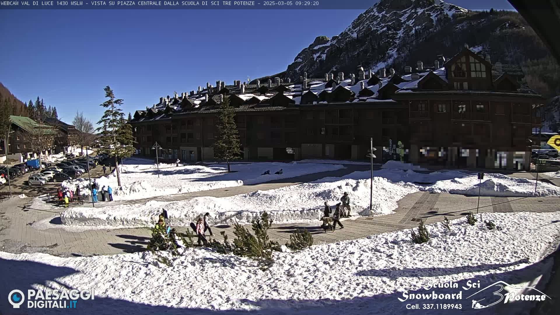 A sunny day in a snow-covered mountain town square, with a large building and several people walking around.