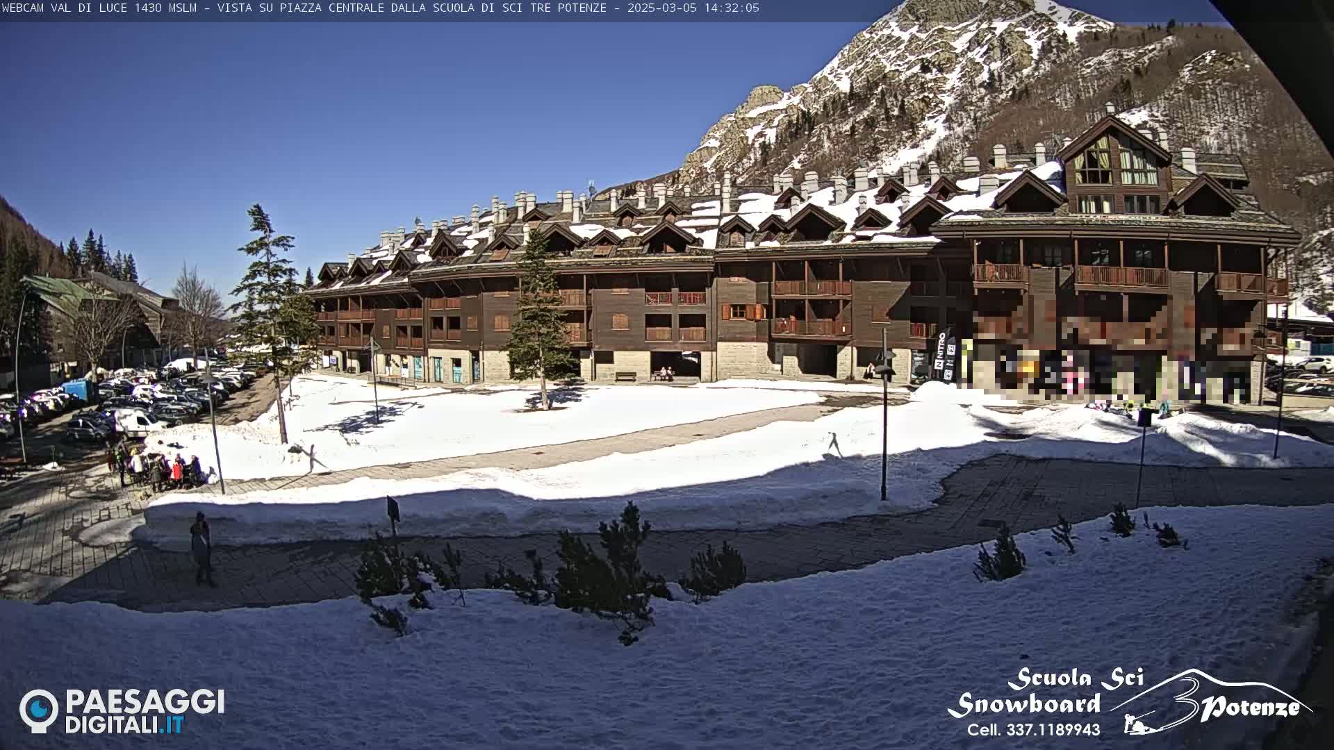 A snow-covered plaza in front of a multi-story brown building, situated against a sunny, mountainous backdrop.