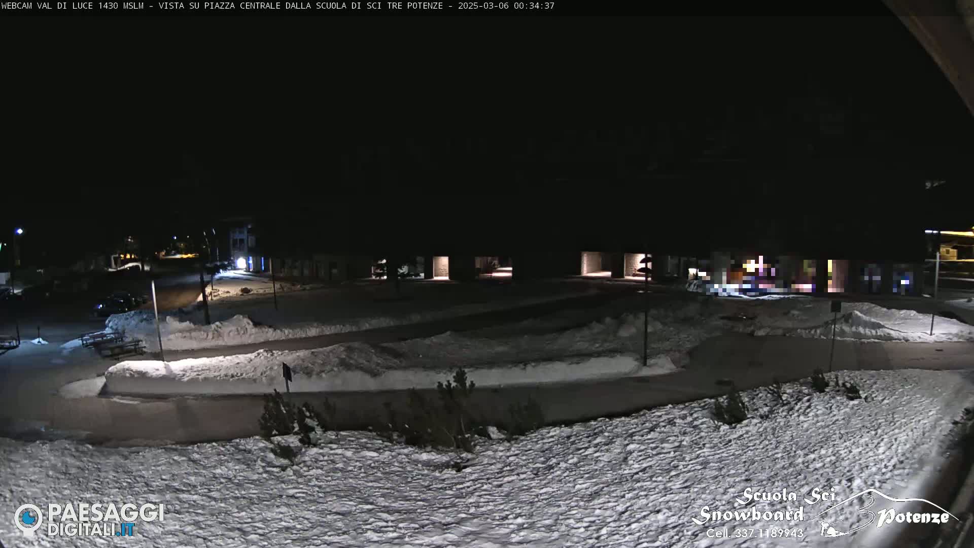 A snow-covered plaza at night, illuminated by dim lights, with a few parked cars visible in the distance.