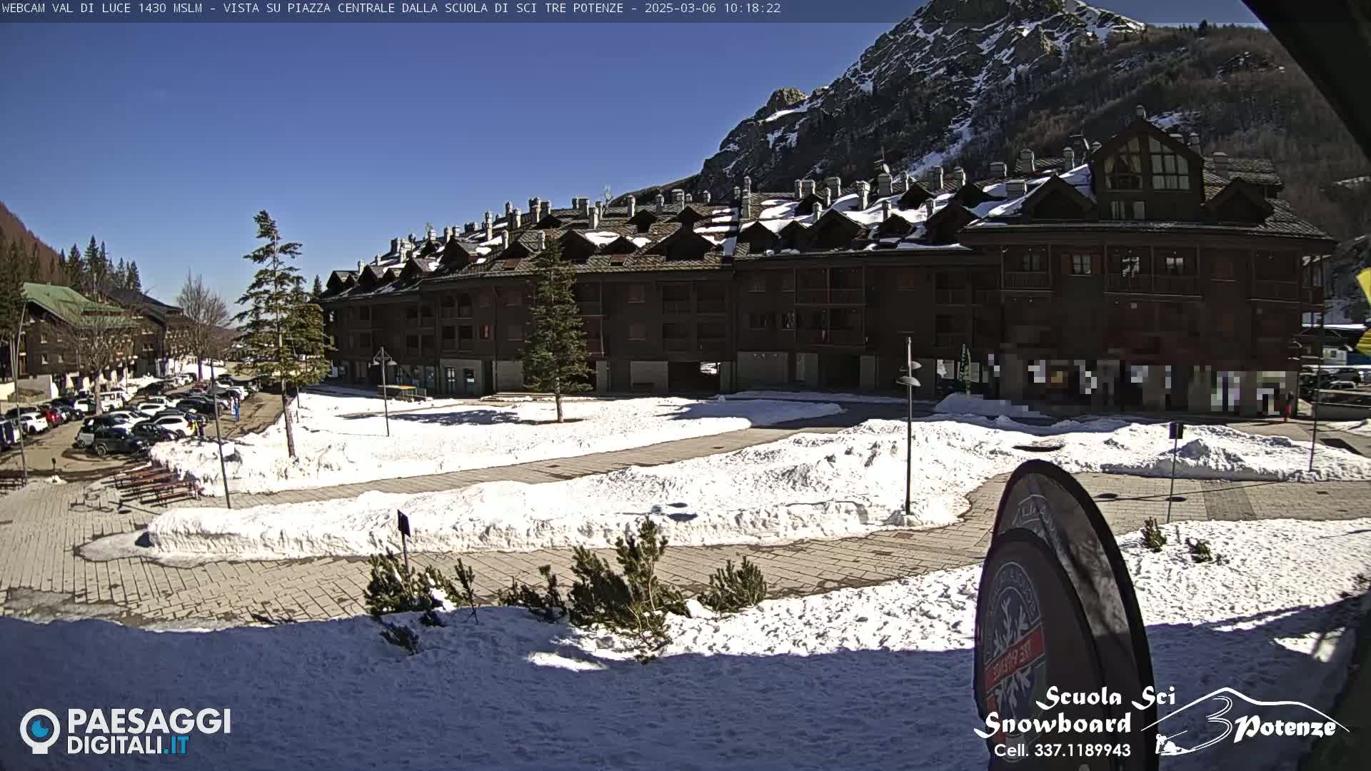 A snow-covered plaza in front of a multi-story building, situated in a mountainous area under a clear sunny sky.