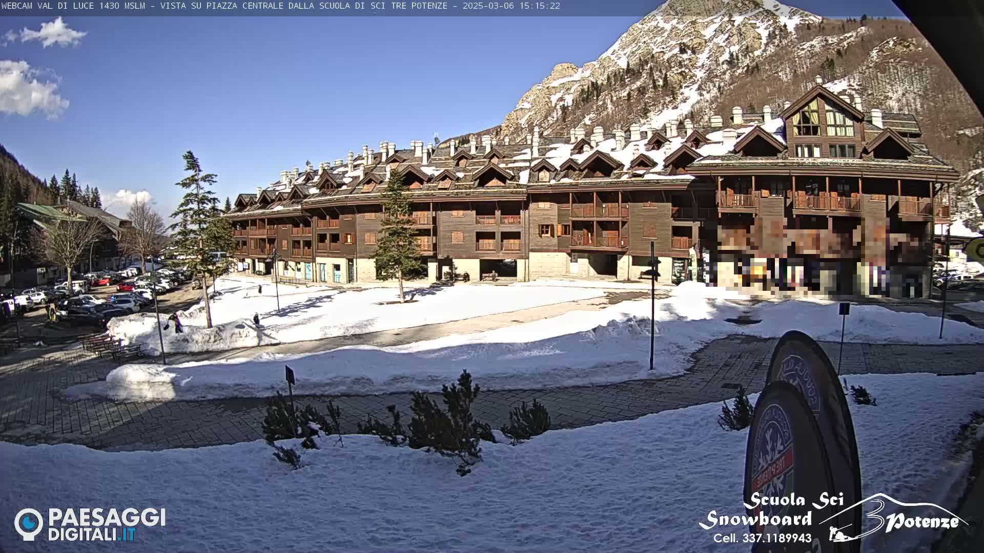 A snow-covered plaza sits in front of a large, multi-story wooden building, with mountains and a partly sunny sky in the background.