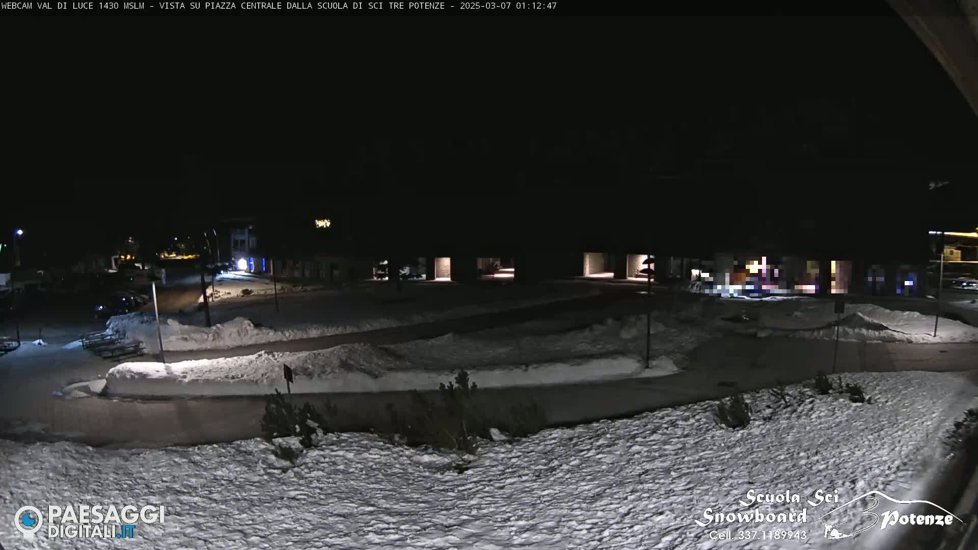 A snow-covered plaza at night, illuminated by sparse lights, with some vehicles parked nearby.