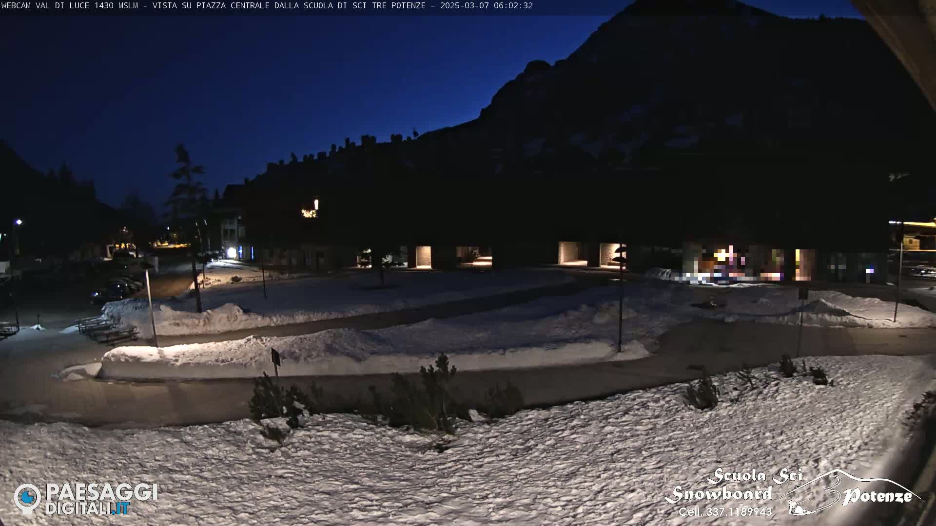 A snow-covered plaza at night, illuminated by a few lights, is nestled beneath a dark mountain under a twilight sky.